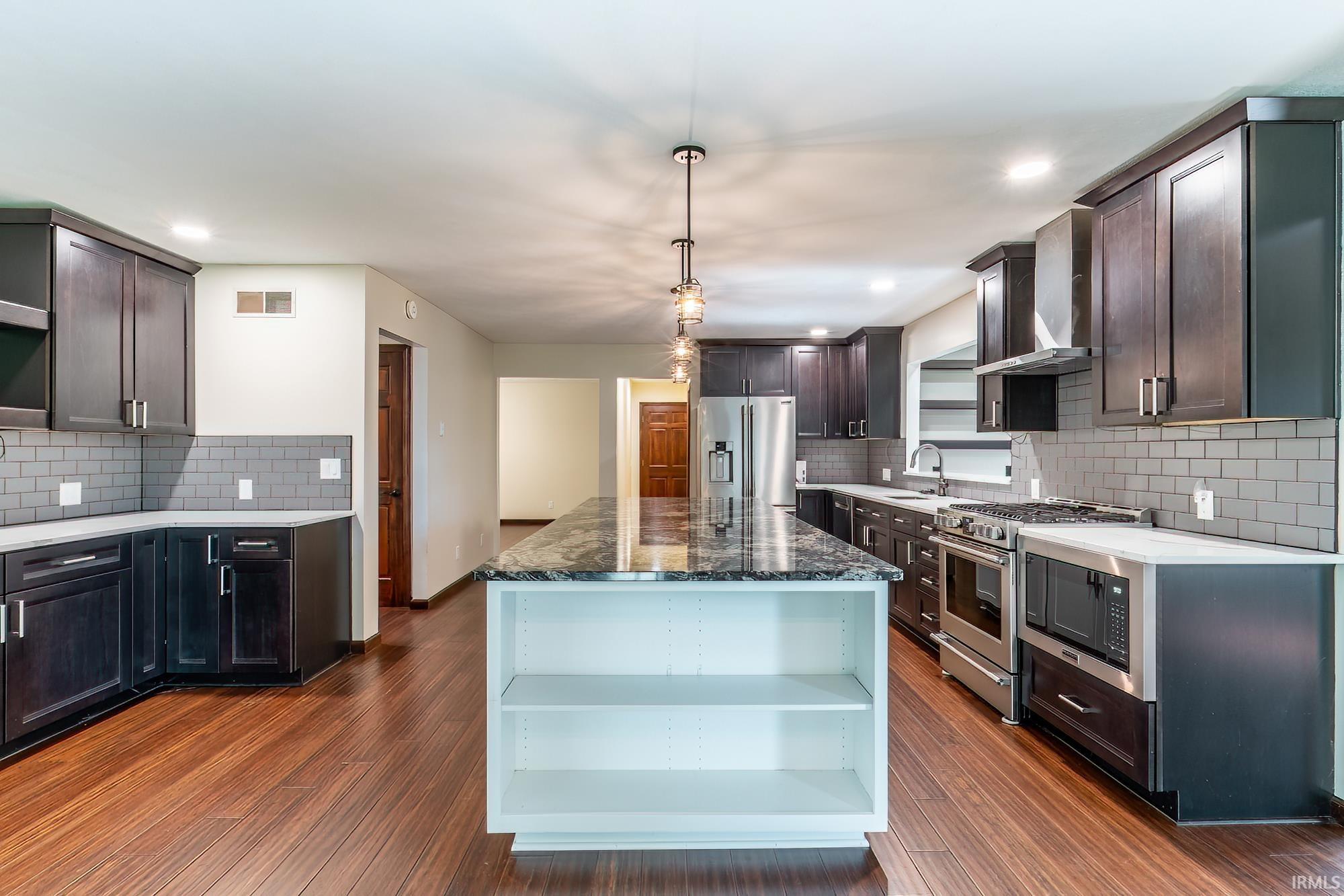 Kitchen with a large granite island, dark cabinets, stainless steel appliances, and wood-look flooring.