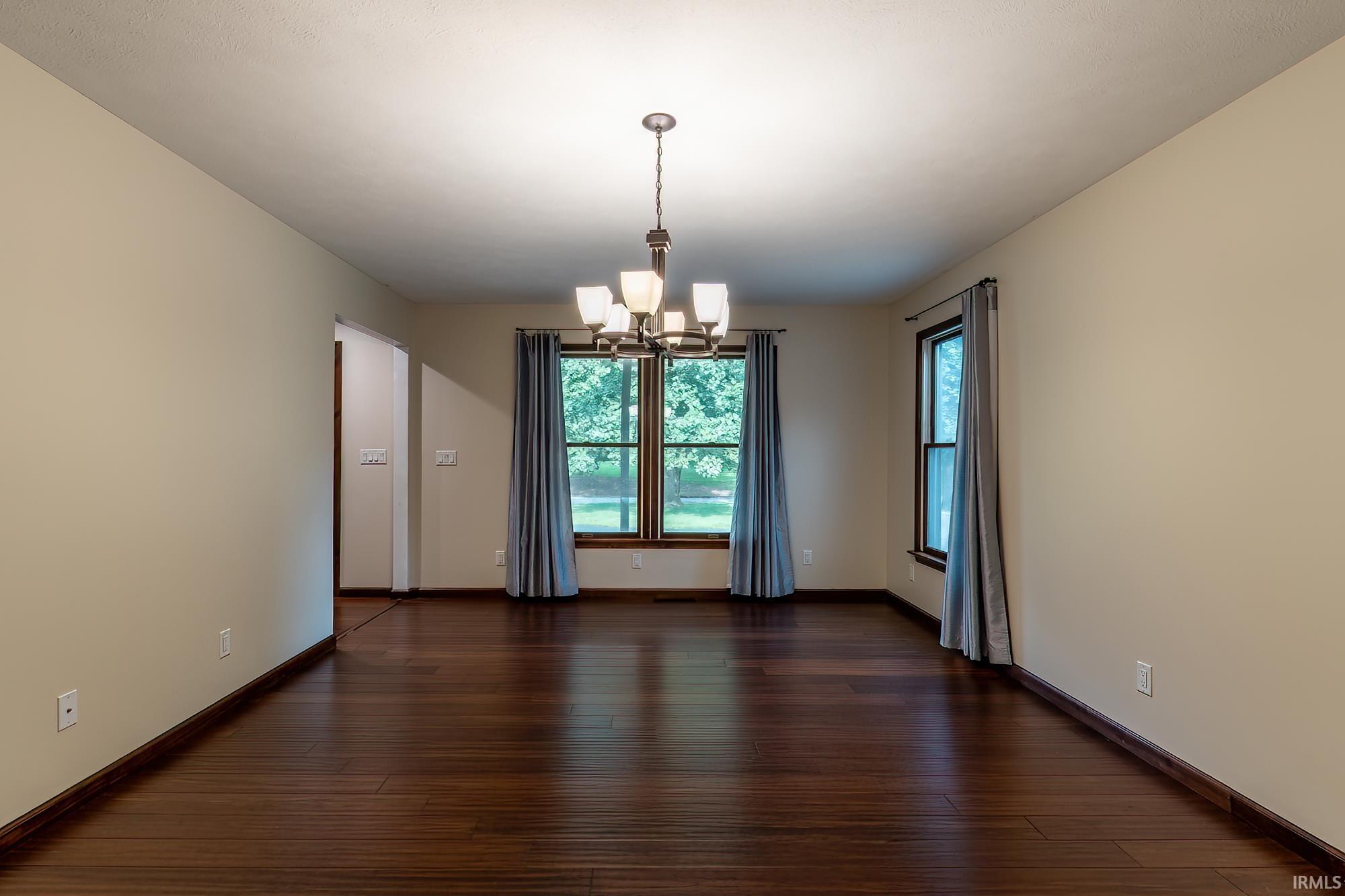 Empty dining room with dark hardwood floors, a chandelier, and curtained windows overlooking greenery.