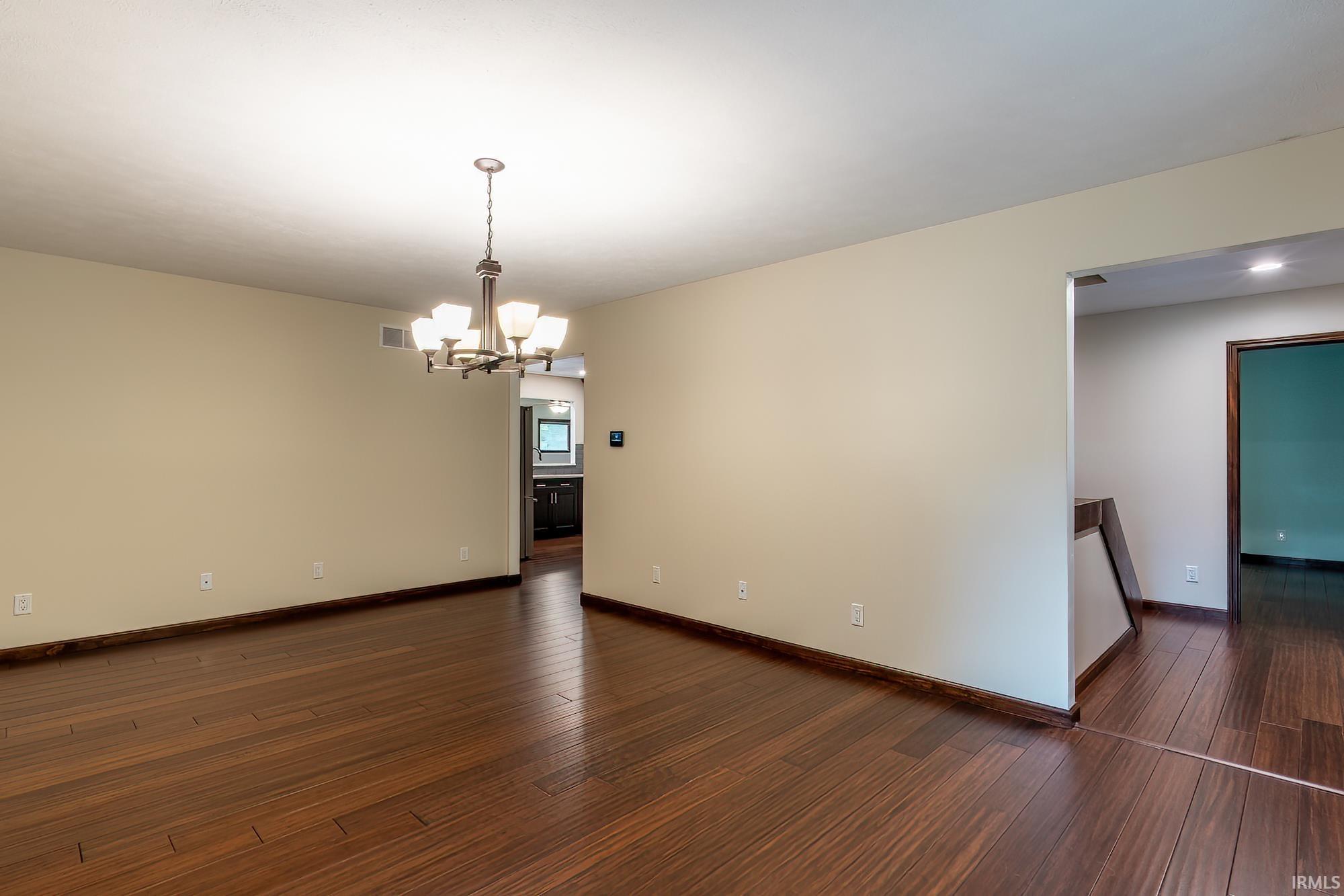 Empty dining room with dark hardwood floors and a lit chandelier, viewed toward adjoining rooms.