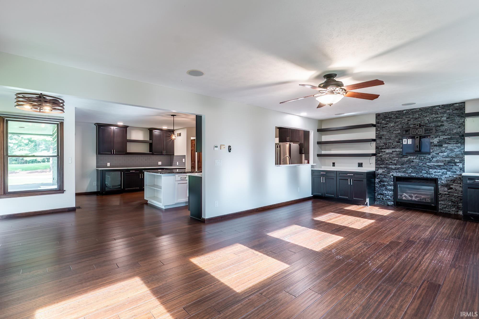 Open living room with dark wood floors, a stone fireplace wall, and kitchen cabinetry.