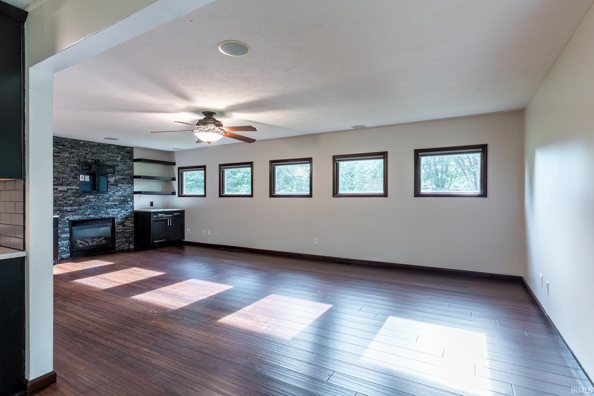 Vacant living room with a stone fireplace wall, five windows, and sunlight across dark plank flooring.