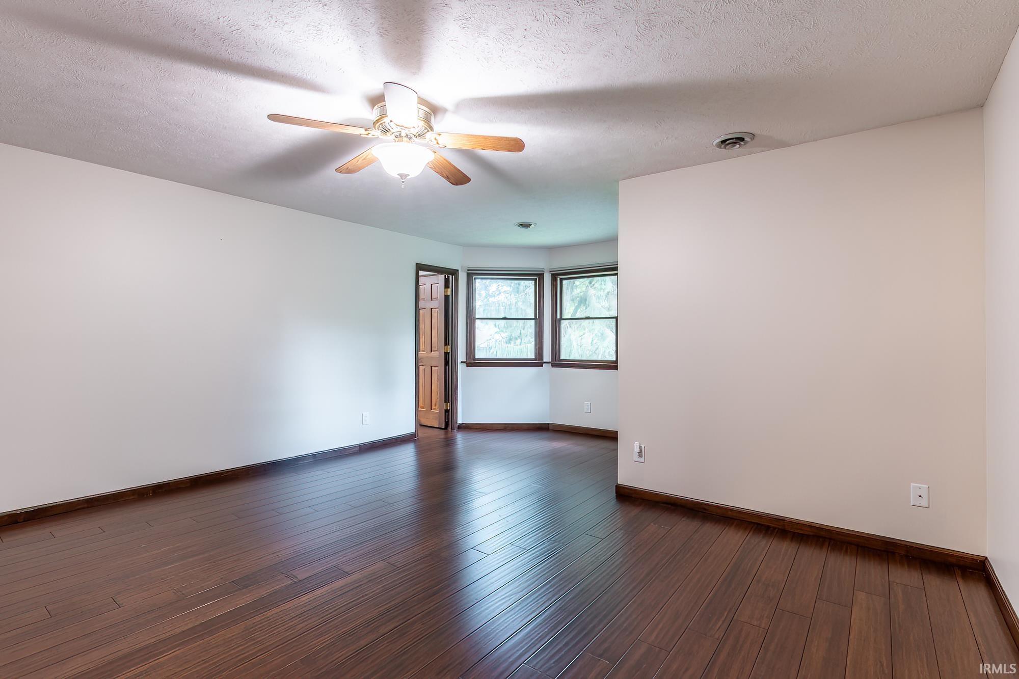 Empty room with dark wood floors, a ceiling fan, and two windows beside an open door.