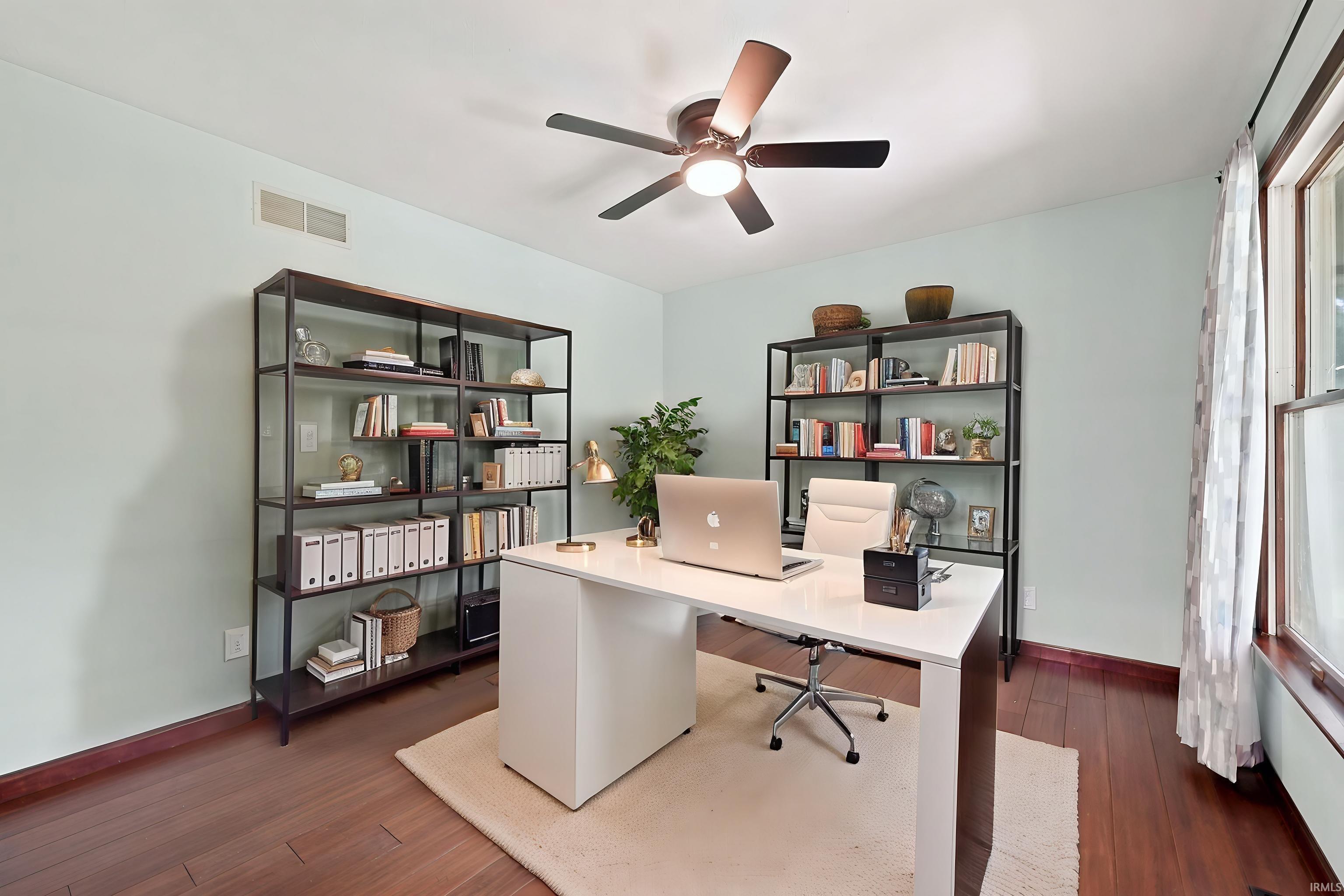 Furnished home office with a white desk, bookcases, and large windows.