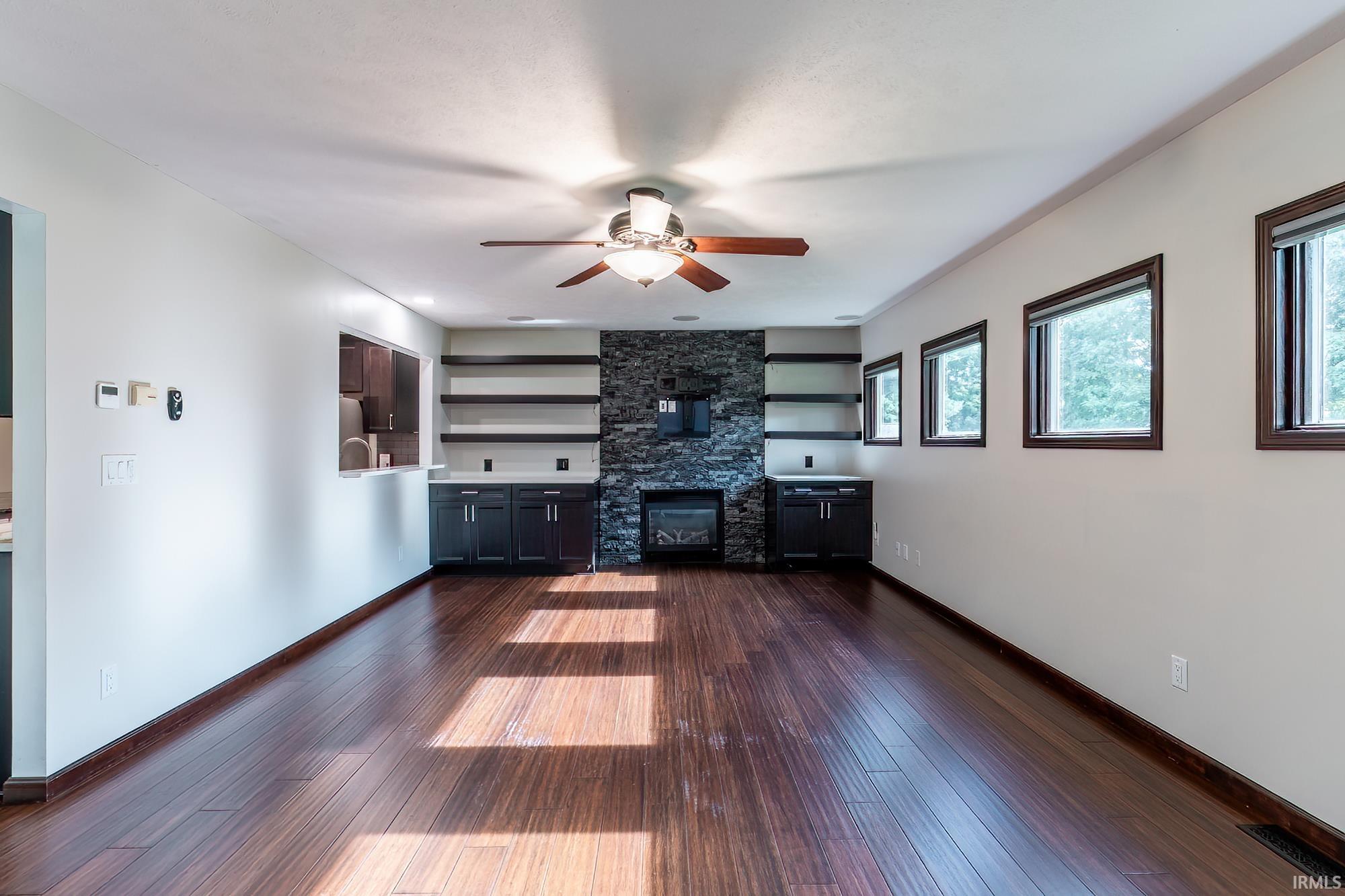 Empty living room with a stone fireplace, built-in shelving, dark flooring, and large windows.