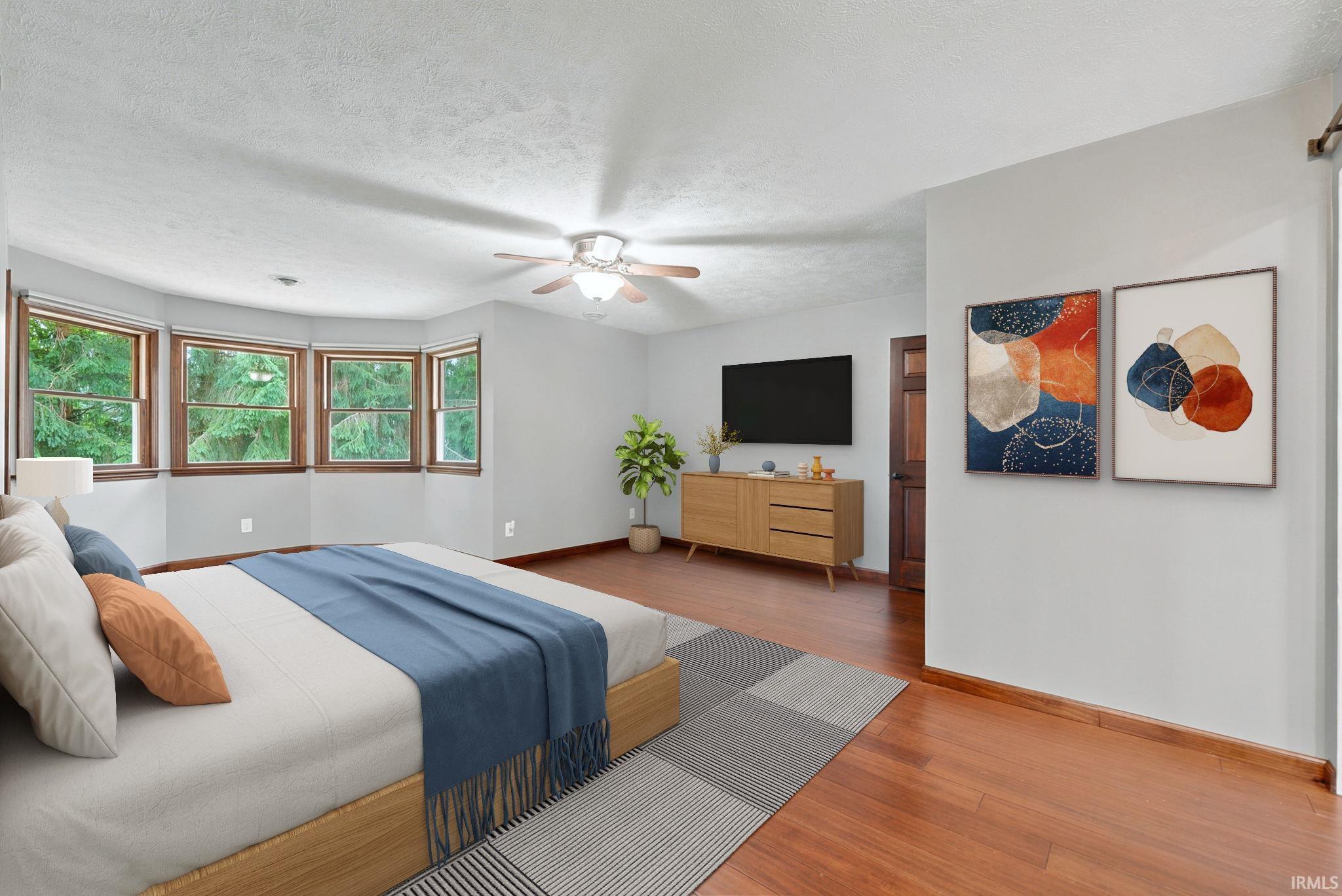 Staged bedroom with wood-trimmed bay windows, blue bedding, and a dresser beneath a wall-mounted TV.