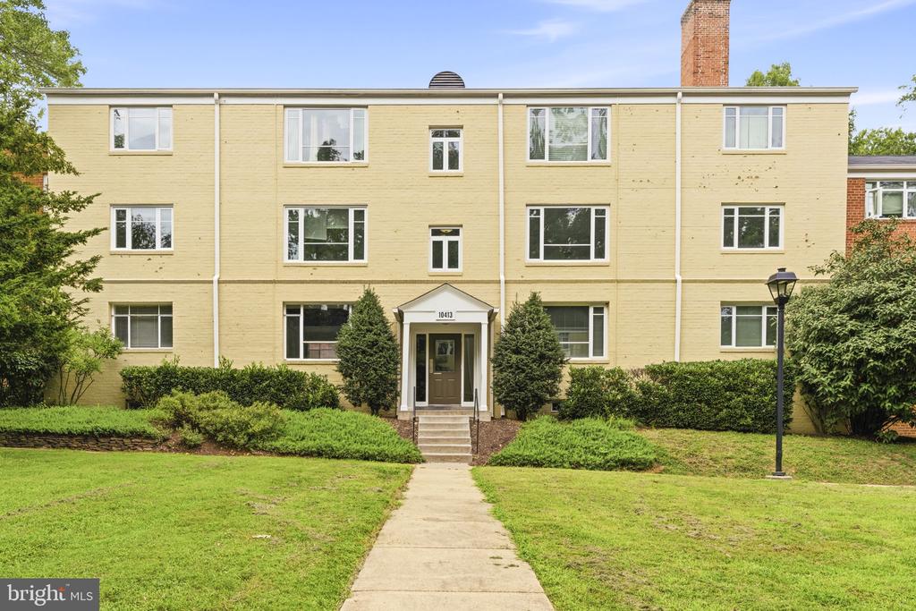 Pale yellow three-story apartment building with a central entrance and walkway through the front lawn.