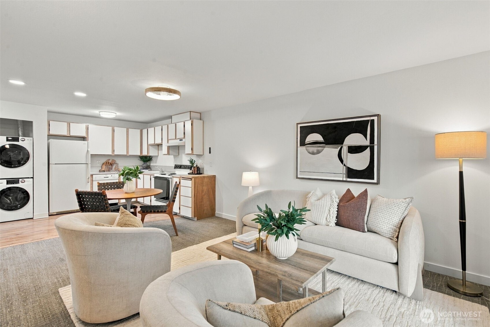 Staged open-plan living room and kitchen with light seating, white cabinets, and a stacked washer and dryer.