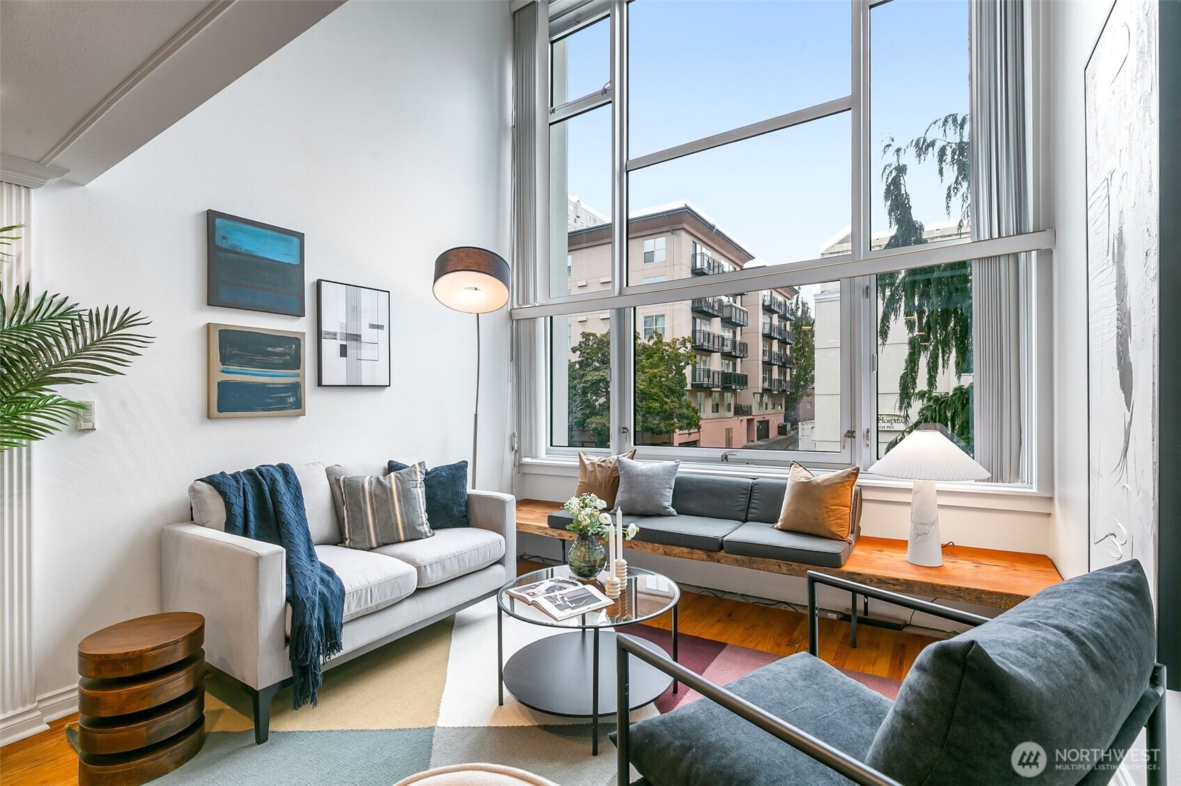 Living room with floor-to-ceiling windows, gray sofas, and views of nearby buildings and trees.