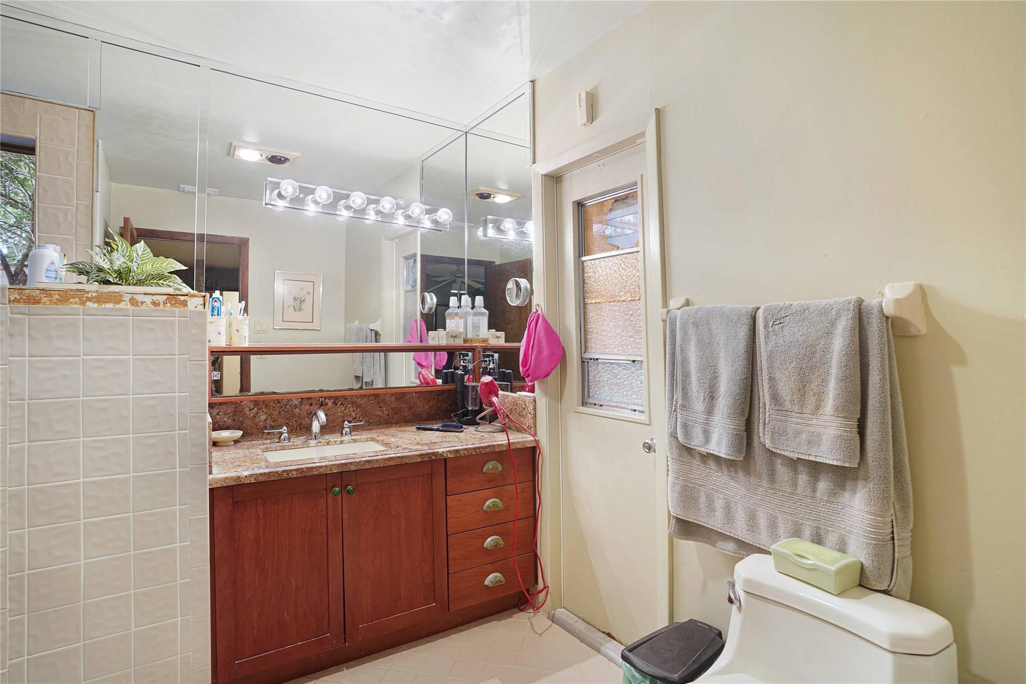 Bathroom with a wood vanity and stone counter beneath a large mirror, beside a tiled shower and frosted window.