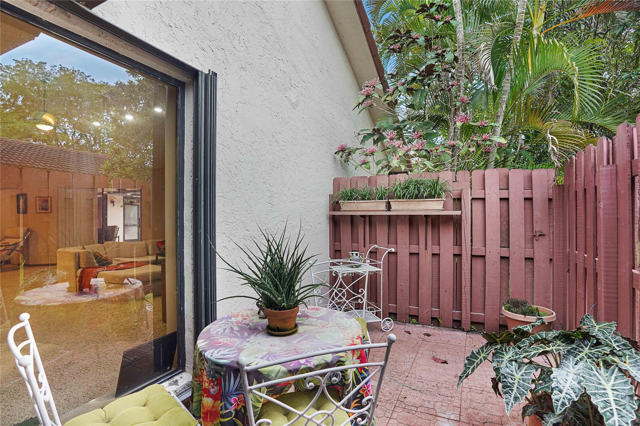 Fenced patio with tropical plants, a floral-covered table, white metal chairs, and a sliding glass door.