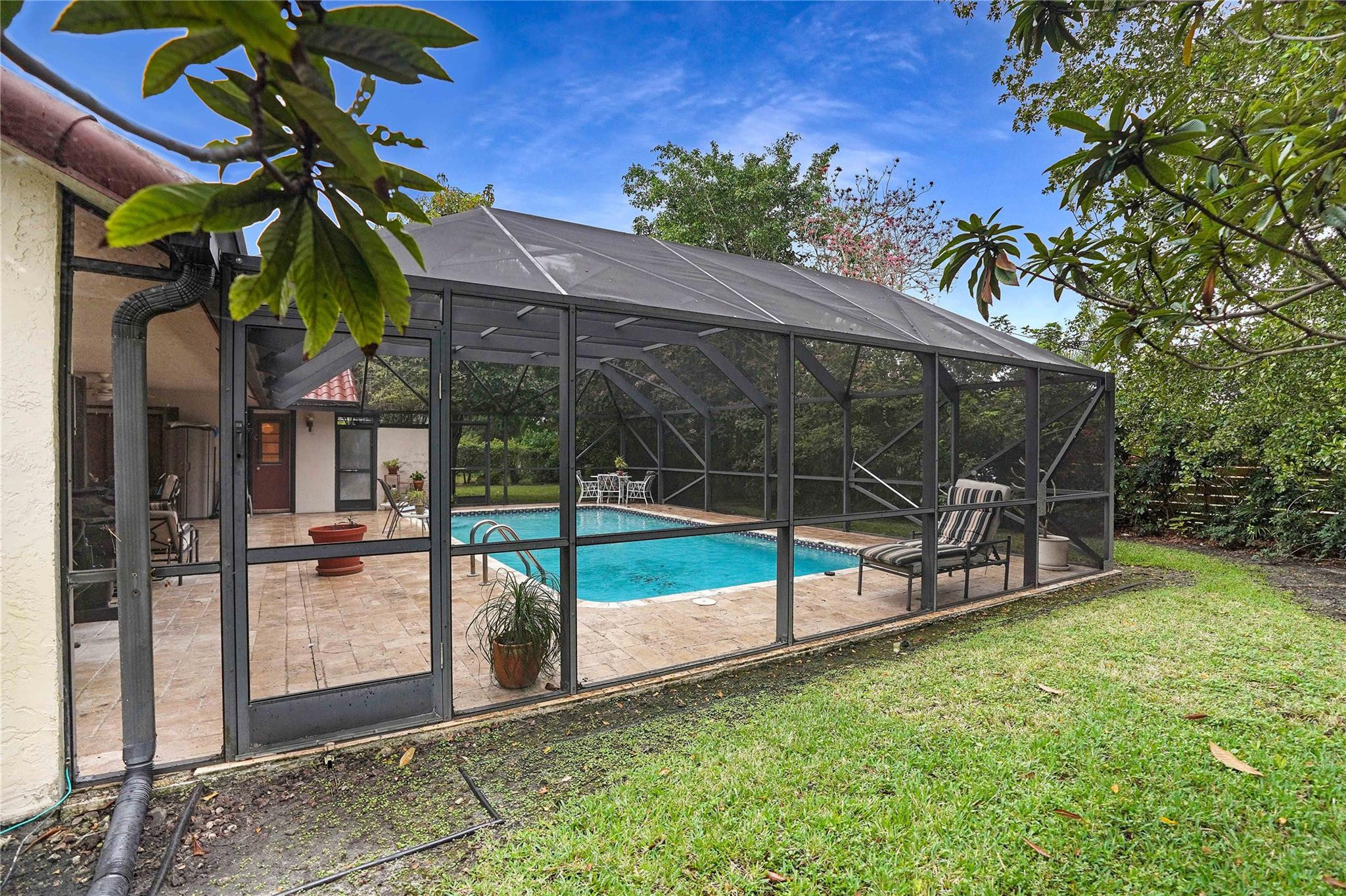 Screen-enclosed backyard pool with a tiled patio, outdoor seating, and surrounding lawn and trees.