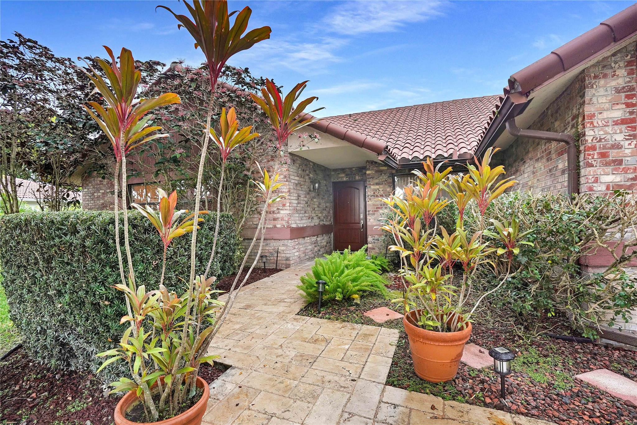 Brick home entrance with a stone walkway, tropical plants, and a red tile roof.