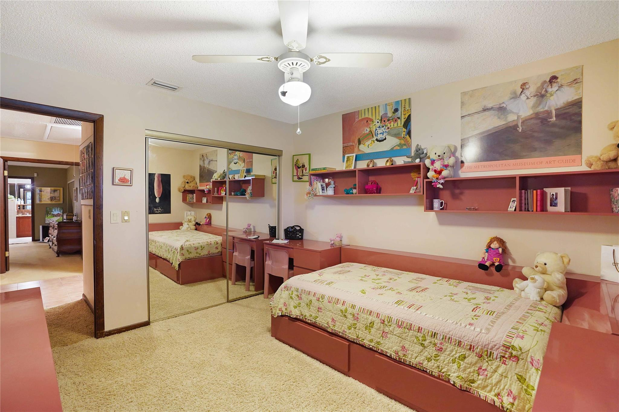 Bedroom with a pink platform bed, floral quilt, mirrored closet doors, and shelves displaying toys.