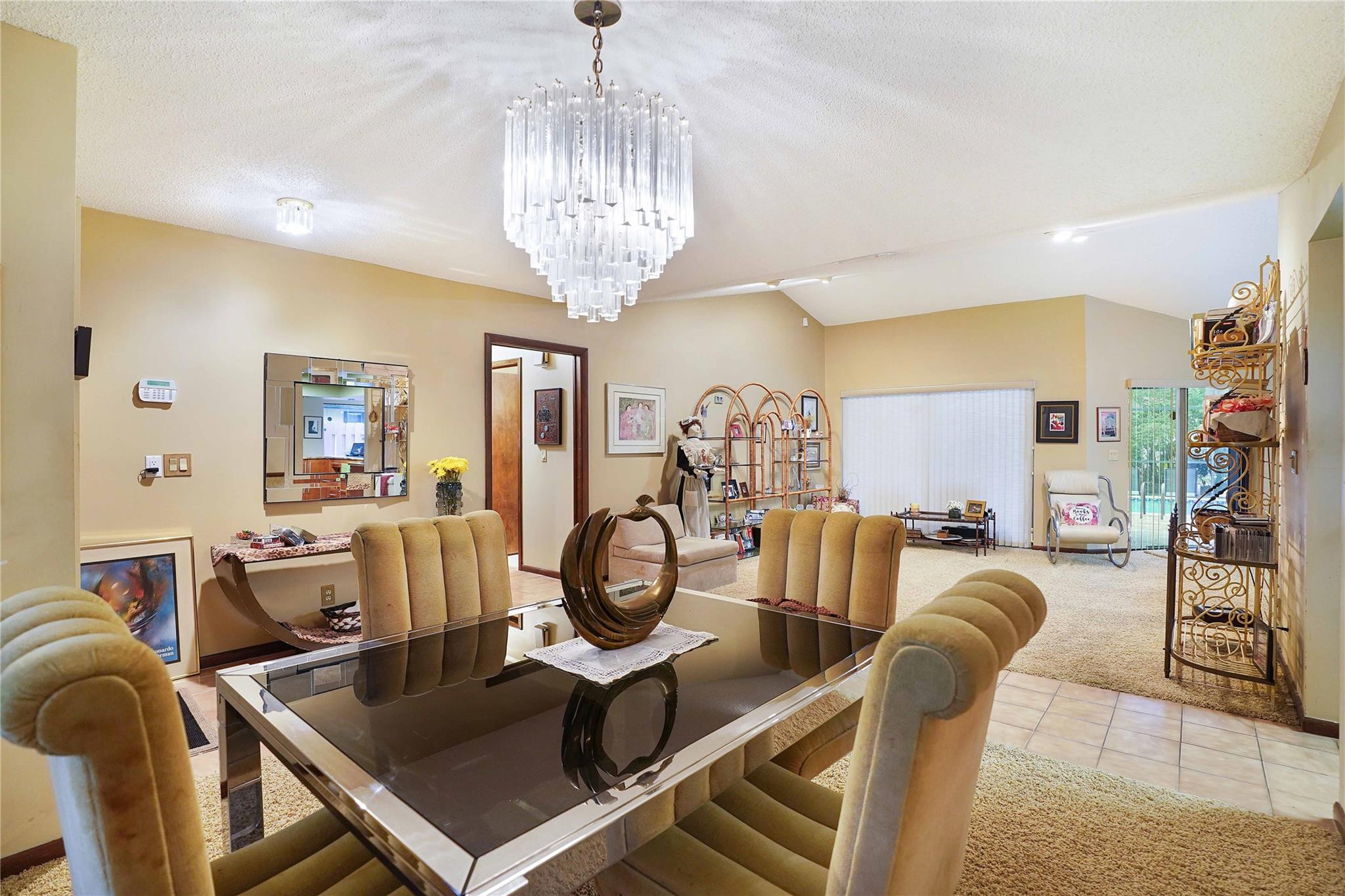 Dining room with a glass table, tan chairs, crystal chandelier, and an open living area.