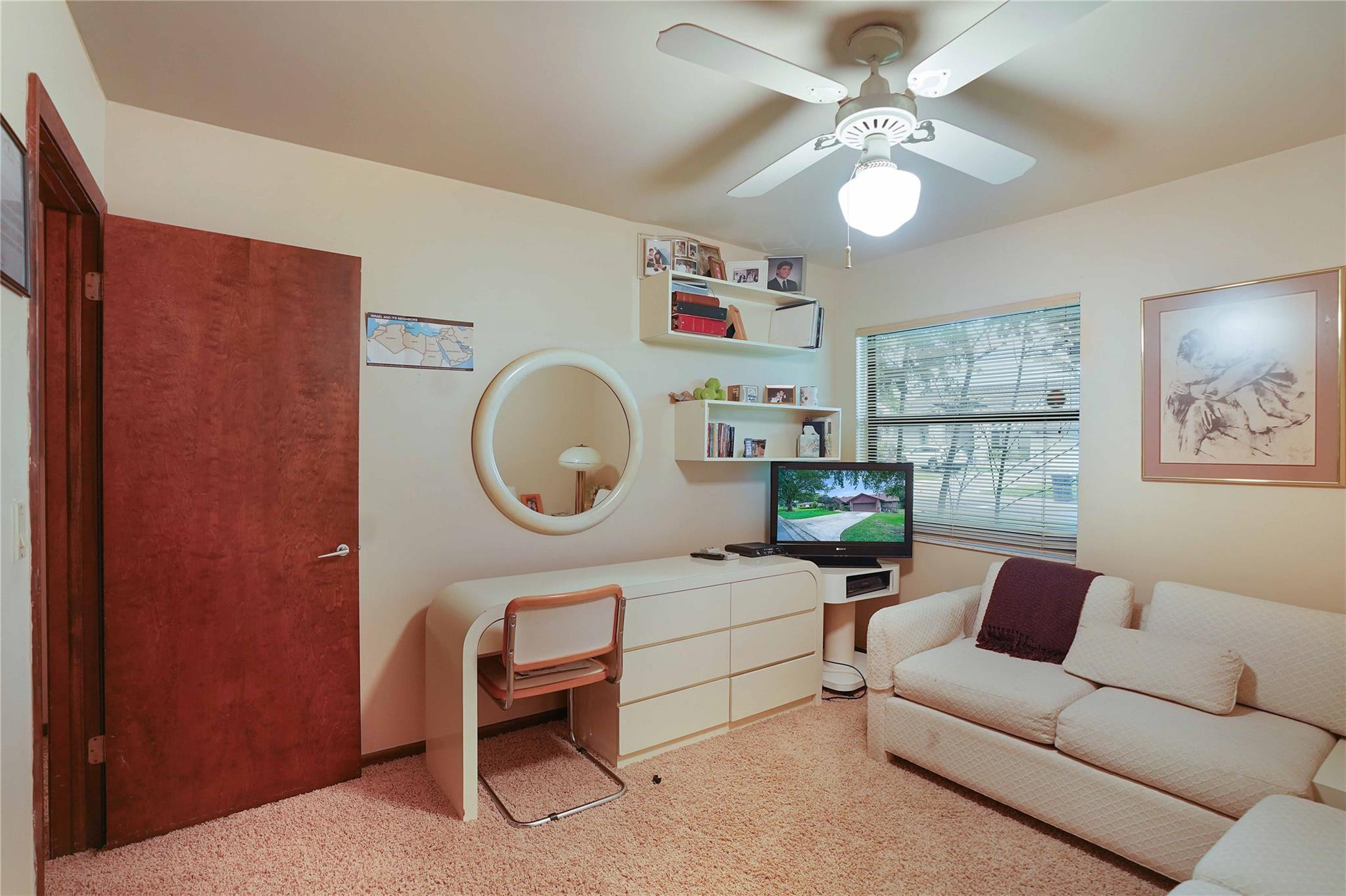 Furnished den with a white sofa, compact desk, wall shelves, and round mirror.