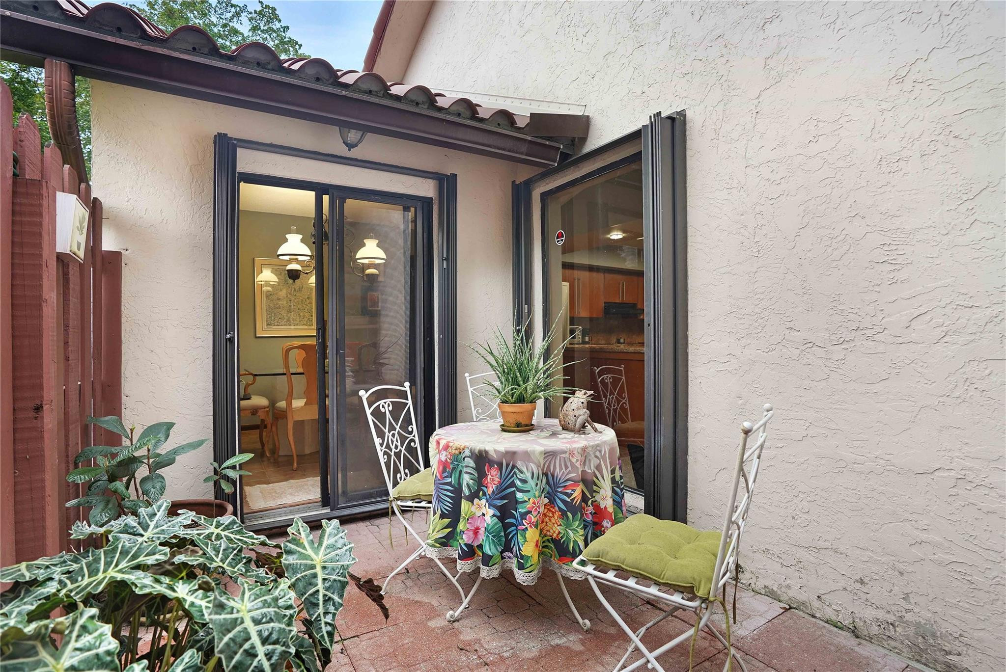Paved patio with a floral-covered table, white chairs, potted plants, and open sliding glass doors.