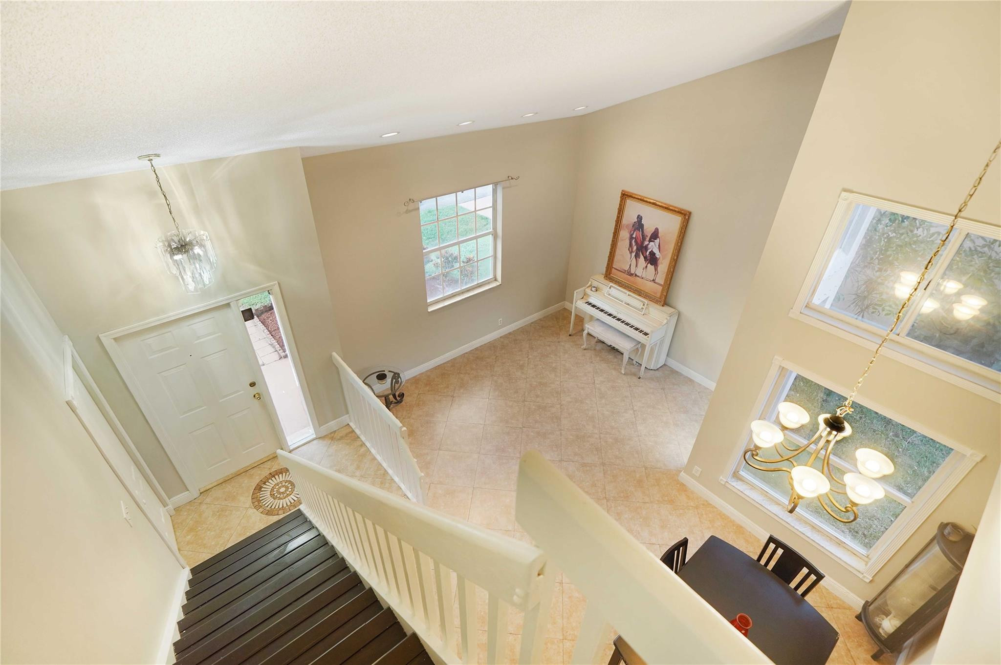 Two-story tiled foyer with a dark staircase, tall windows, chandeliers, and a white piano.