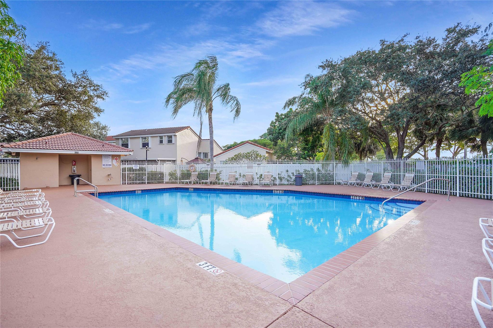 Outdoor community swimming pool with lounge chairs, a white fence, and palm trees.