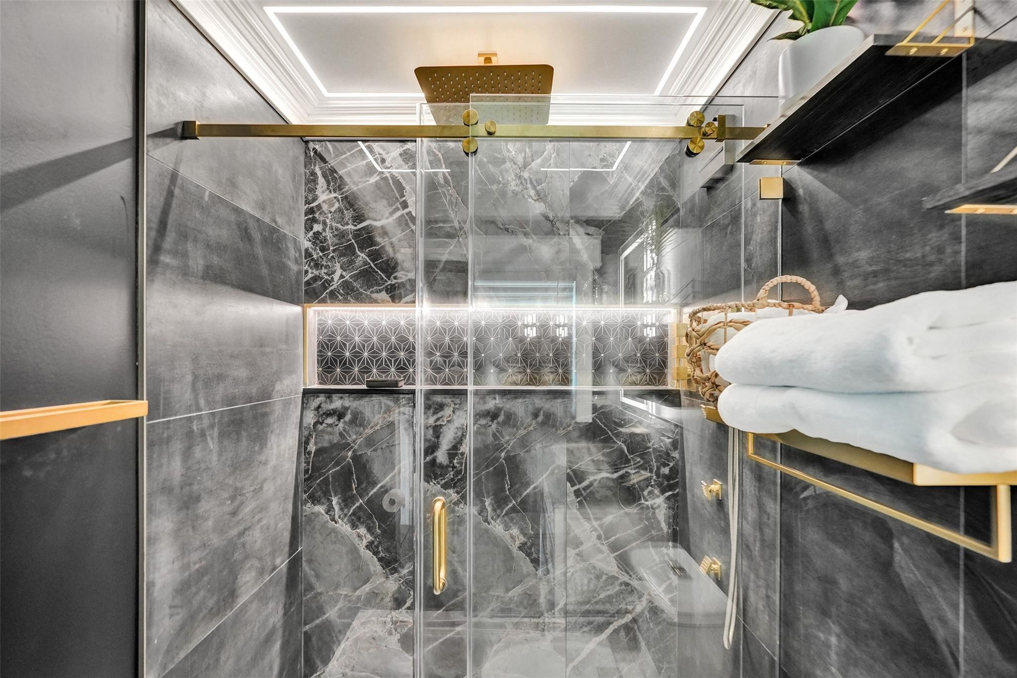 Marble-tiled shower with glass enclosure, gold fixtures, rainfall showerhead, and folded white towels.