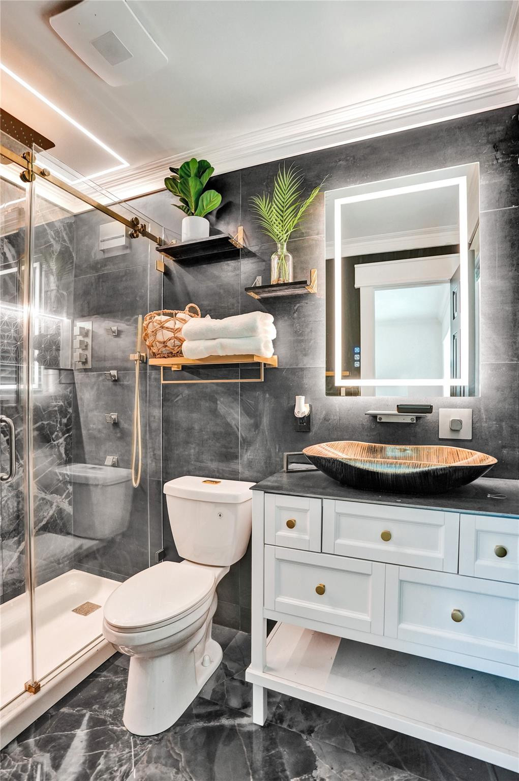 Bathroom with glass shower, white vanity, dark vessel sink, and illuminated mirror.