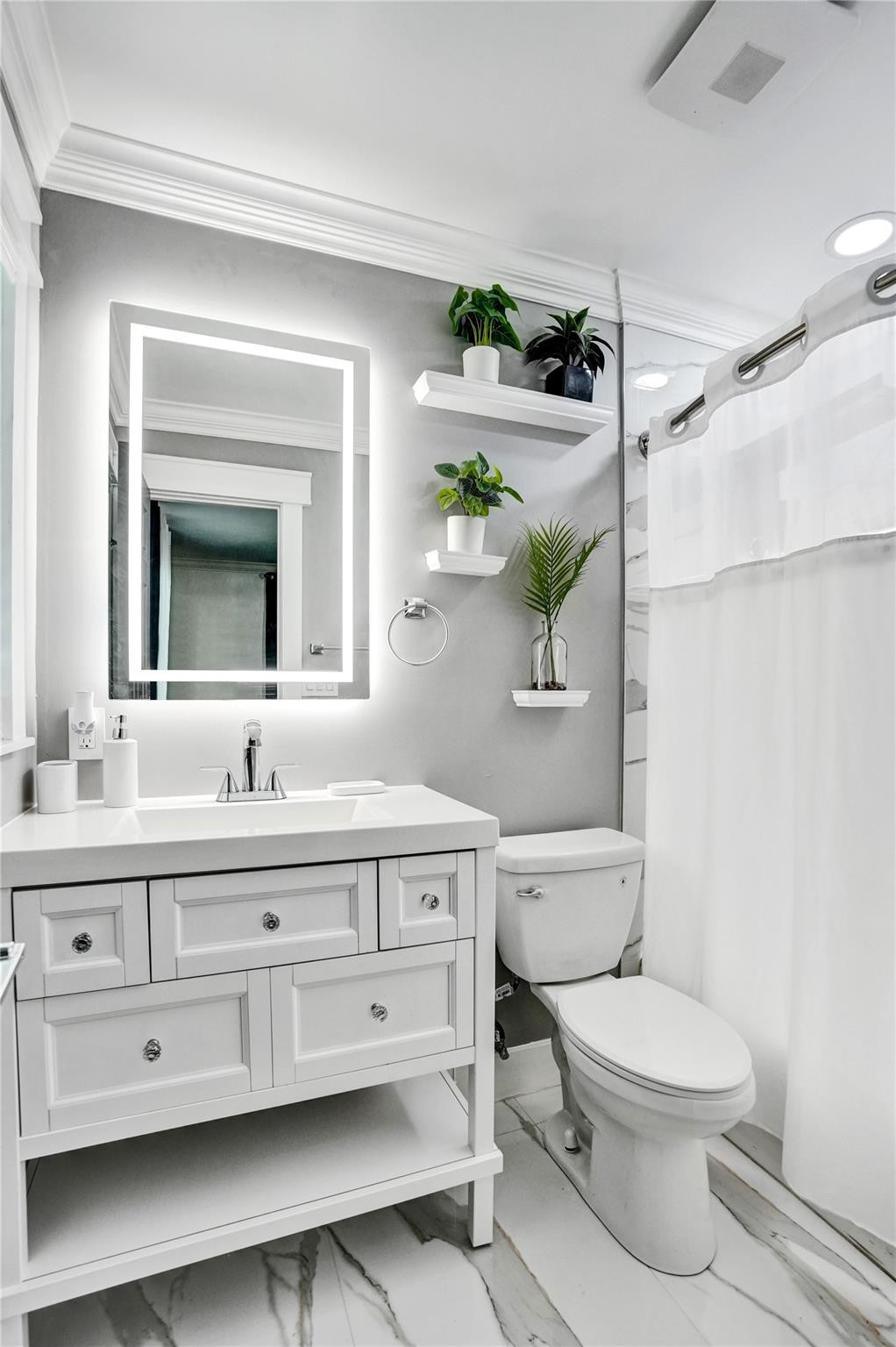 Bathroom with a white vanity, illuminated mirror, marble-pattern floor, and plants on floating shelves.