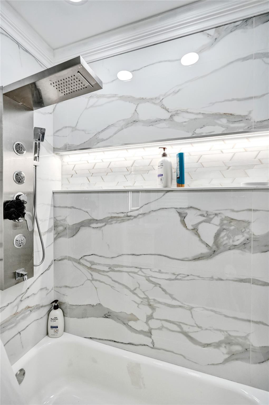 Bathroom tub and shower with marble-look tile, a stainless steel shower panel, and recessed shelf.