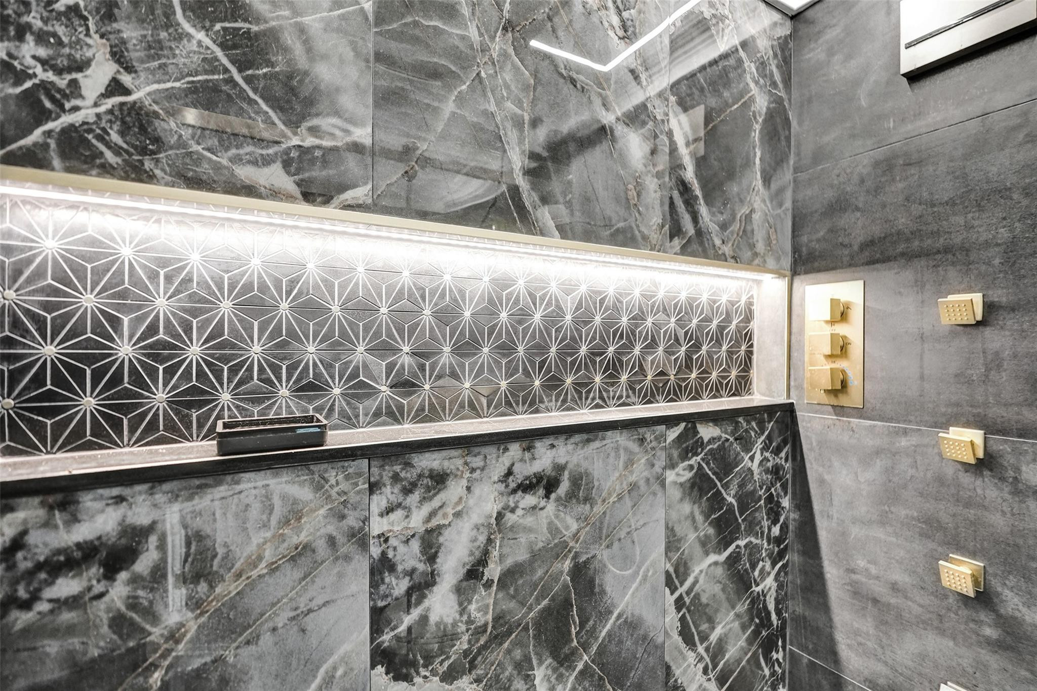 Marble shower wall with illuminated geometric tile and gold-toned controls and body jets.