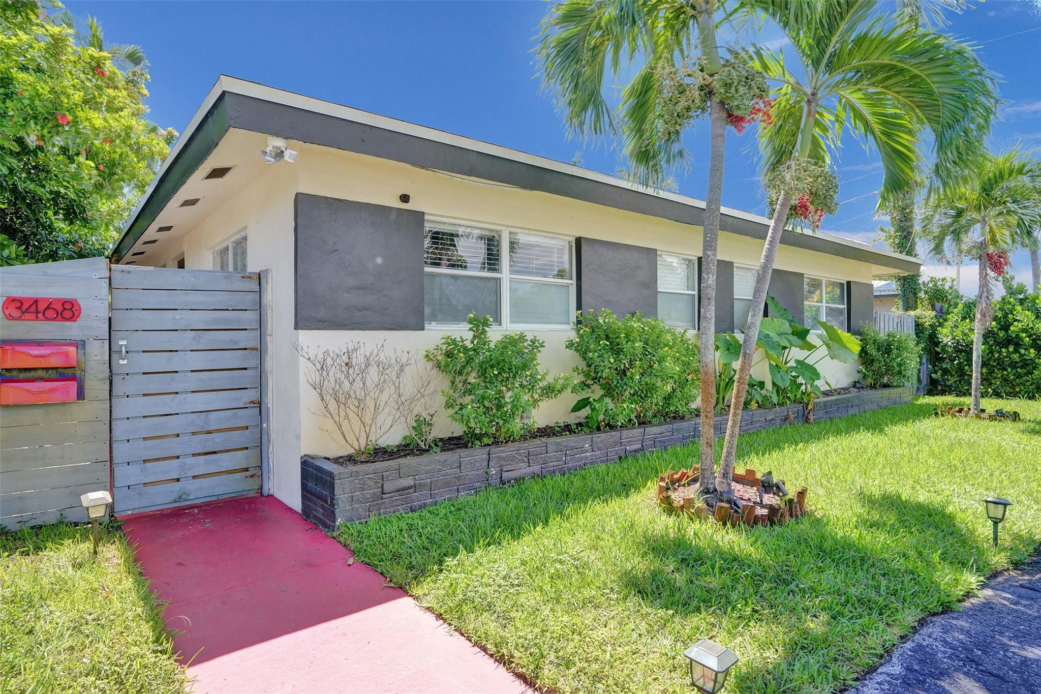 Single-story modern home with a gated entrance, red walkway, and palm trees in the front yard.