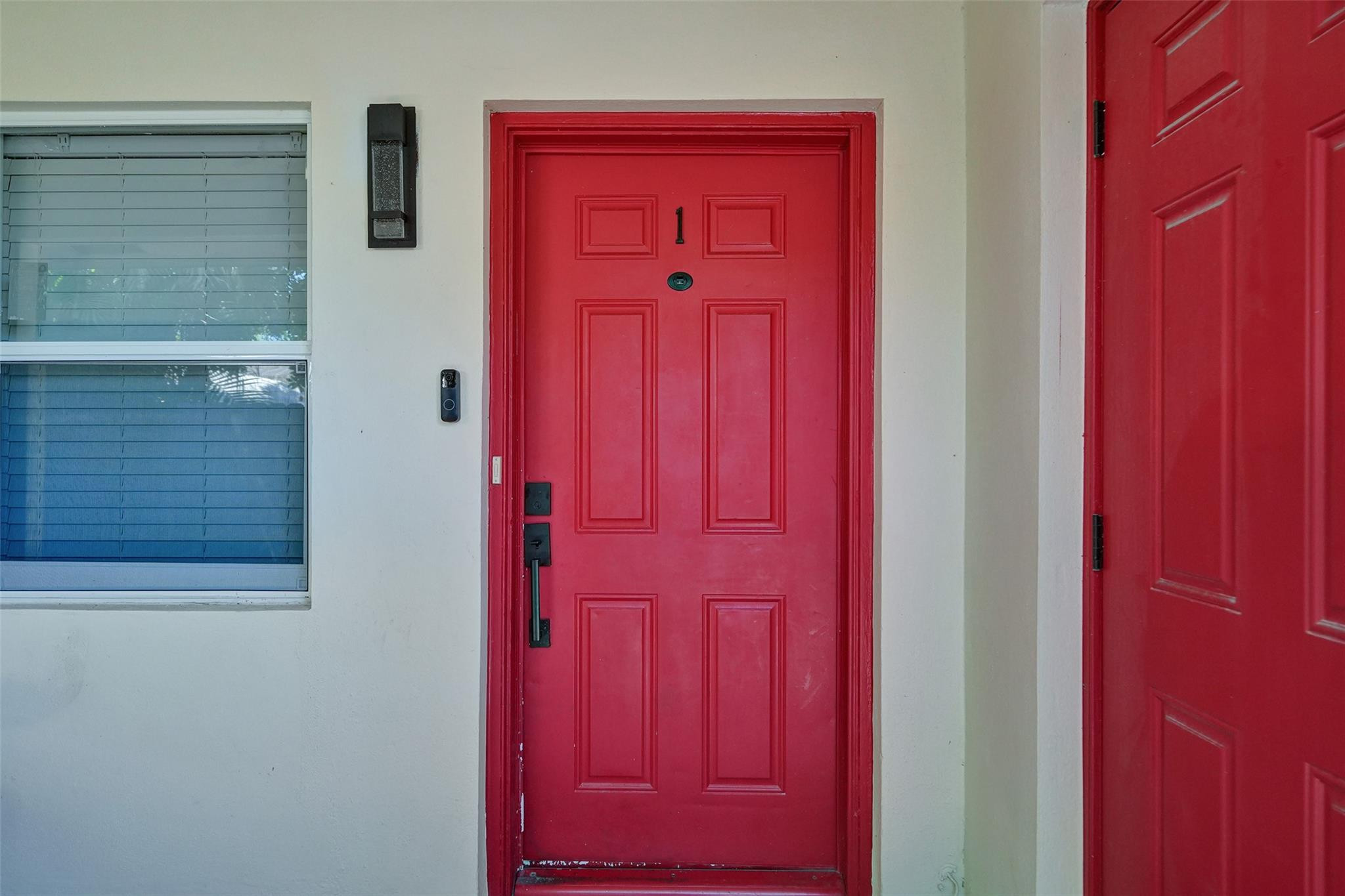 Red paneled front door numbered 1 beside a window with blinds and a video doorbell.