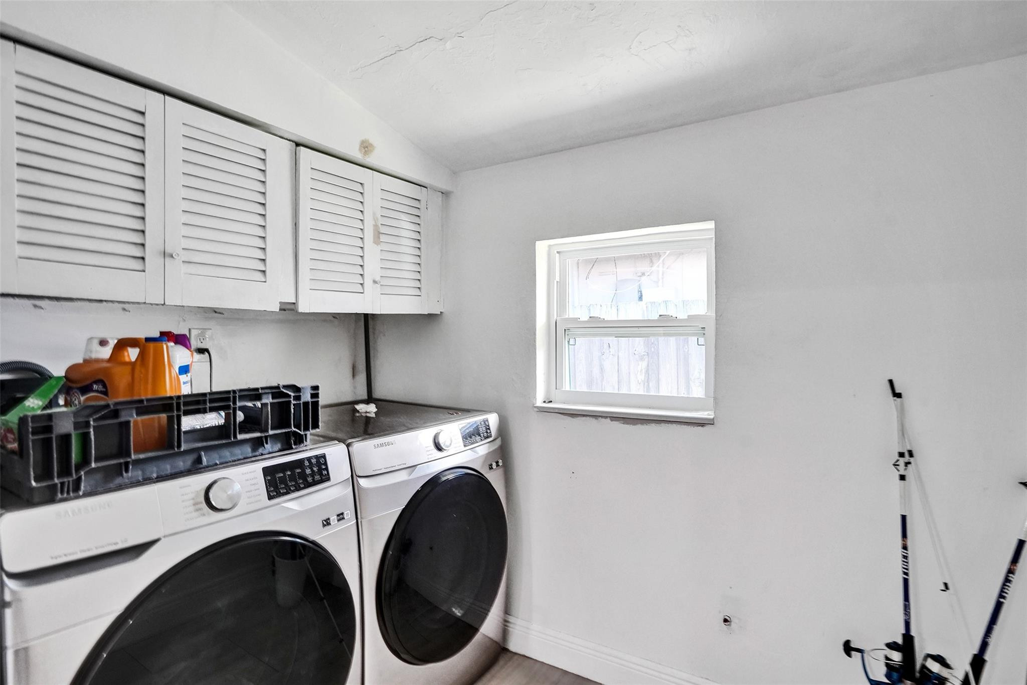 Laundry room with front-loading washer and dryer beneath louvered cabinets, beside a small window.