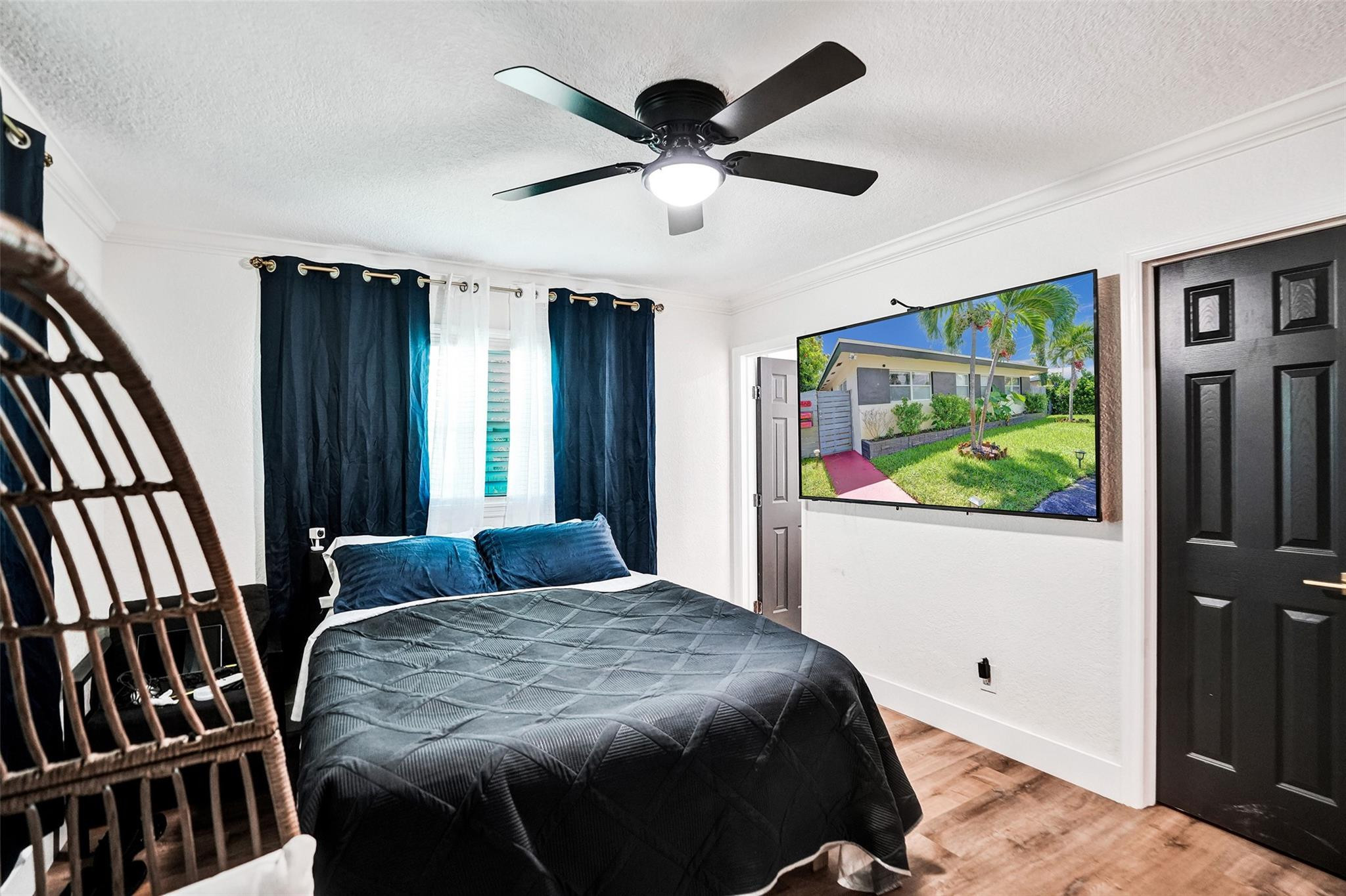 Bedroom with a dark quilted bed, blue curtains, ceiling fan, and wall-mounted television.