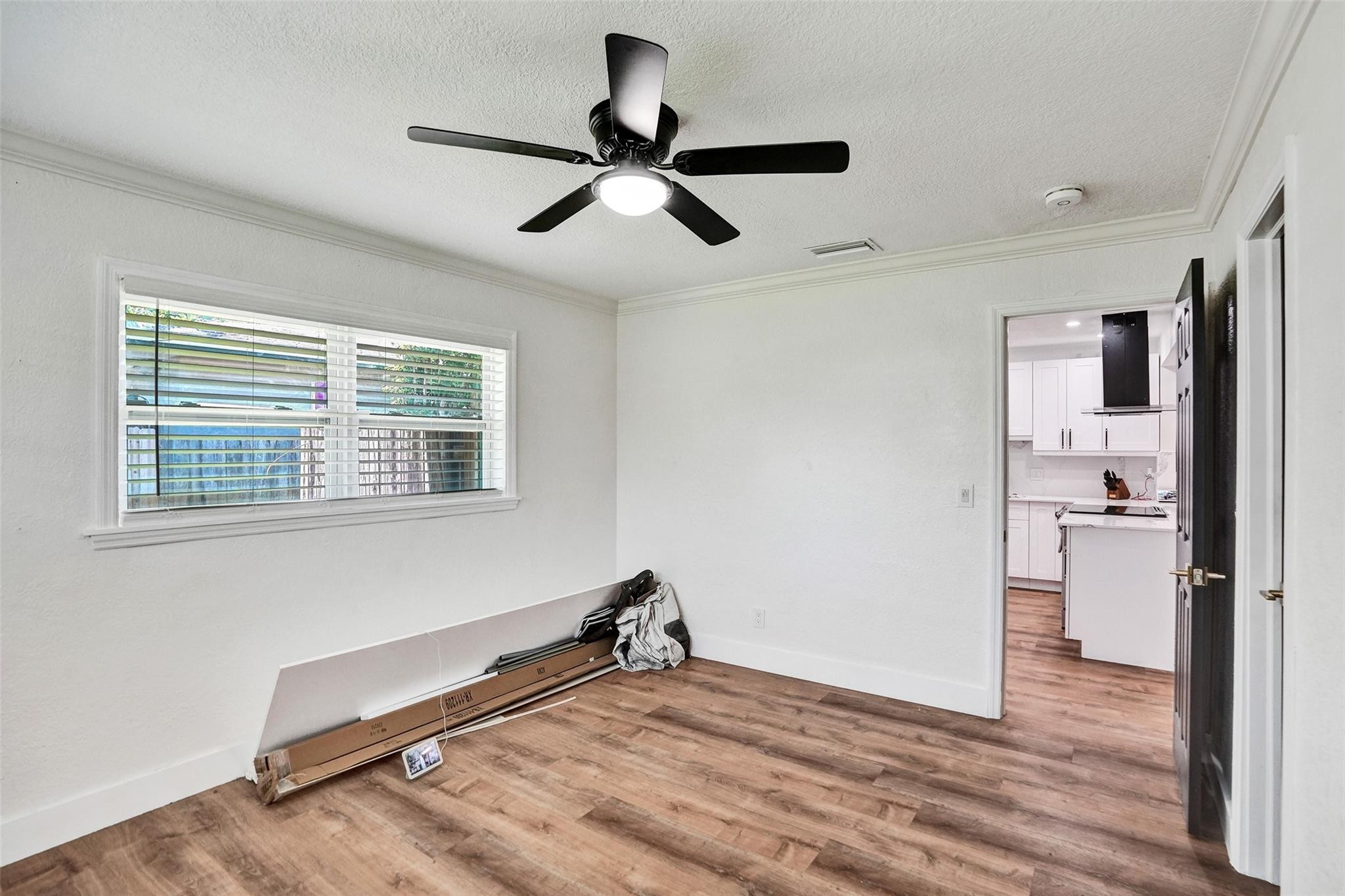 Empty room with wood-look floors and a ceiling fan, opening to a white kitchen.