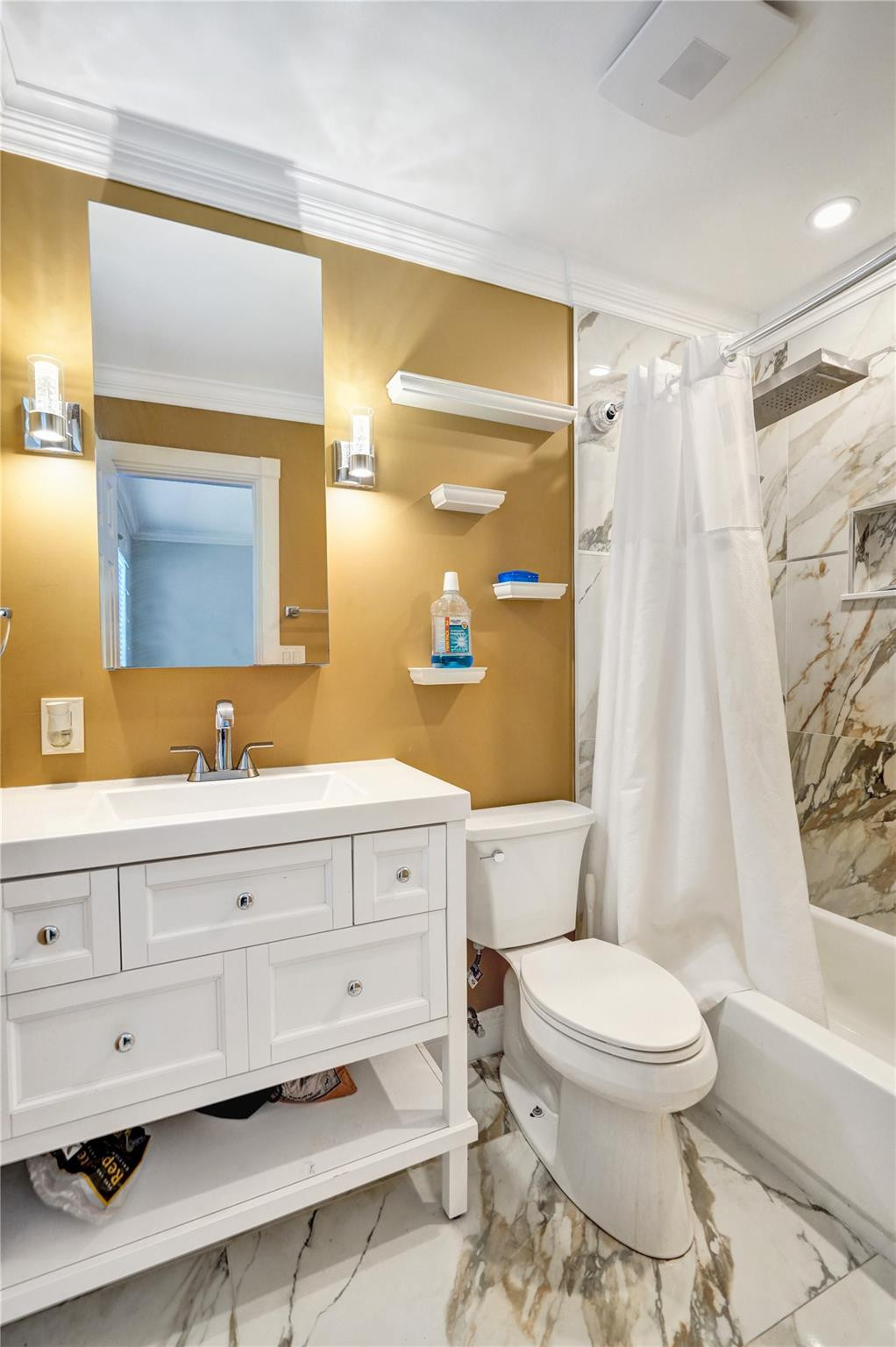 Bathroom with a white vanity, gold walls, and veined marble-look tile around the tub and shower.