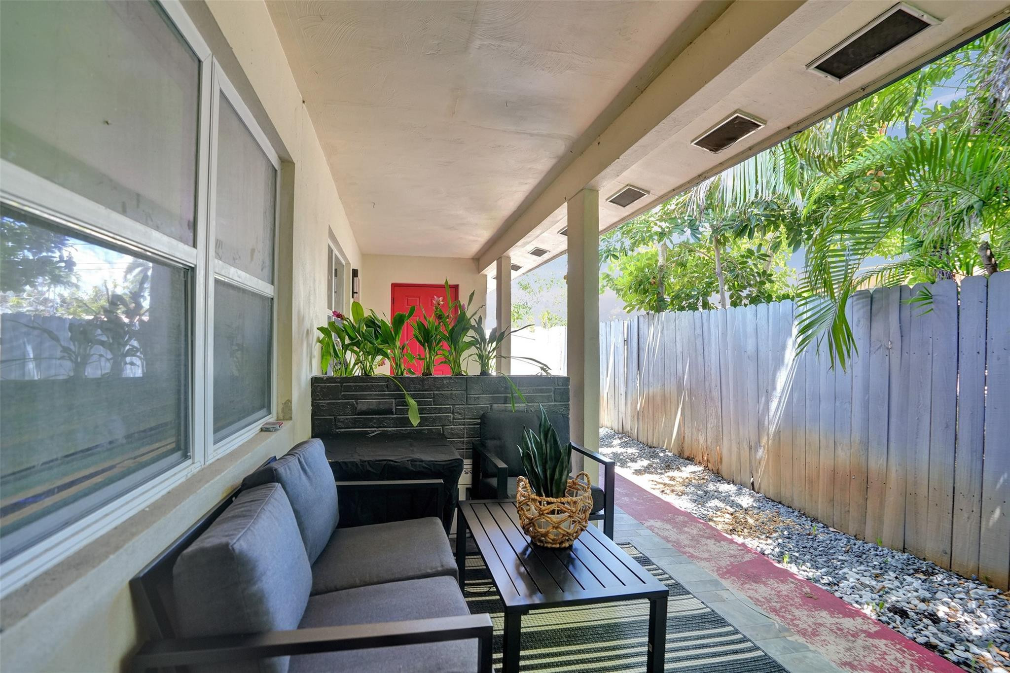 Covered patio with gray seating, a black coffee table, tropical plants, and a wooden privacy fence.