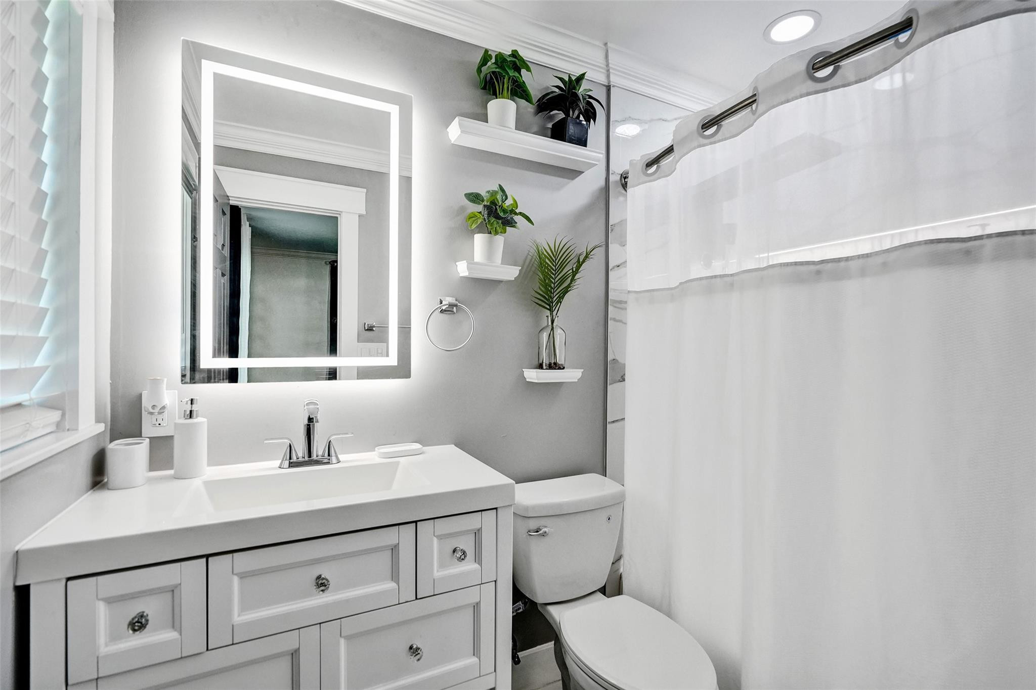 Bathroom with white paneled vanity, illuminated mirror, toilet, and shower curtain; potted plants decorate wall shelves.