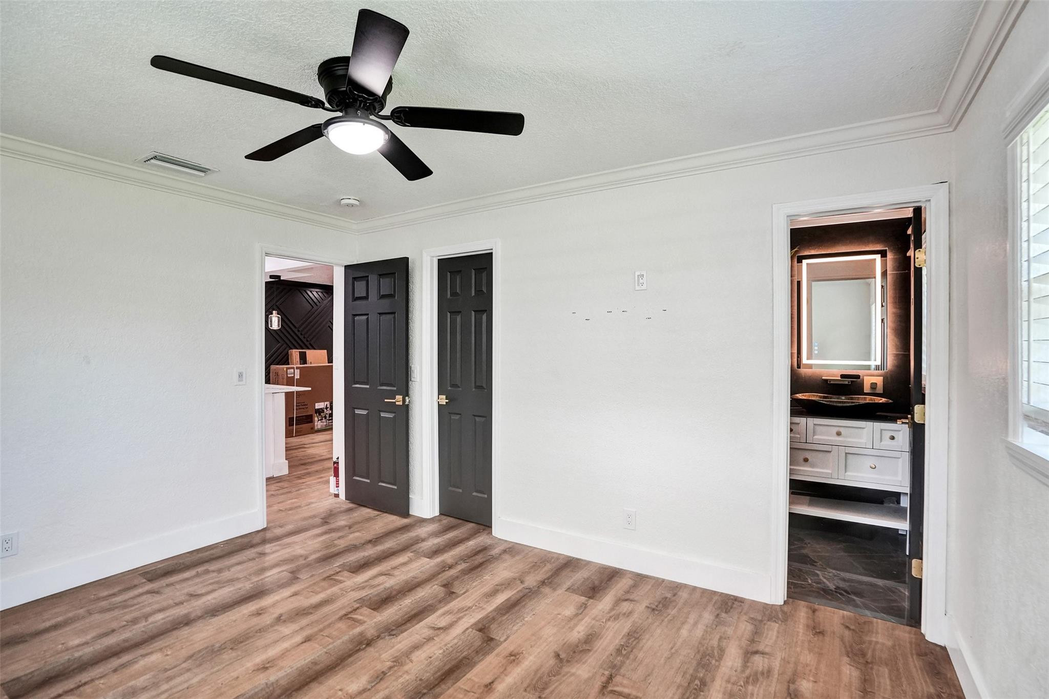 Bedroom with wood-look floors and black ceiling fan, opening to a bathroom with an illuminated mirror.