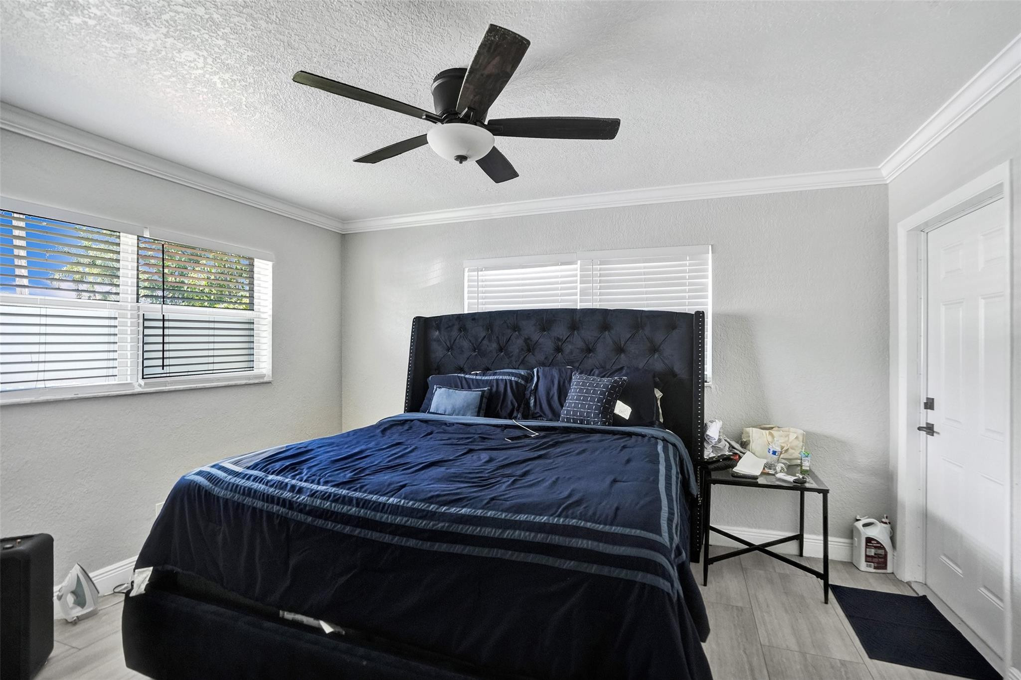 Bedroom with a navy-covered bed, tall tufted headboard, white window blinds, and ceiling fan.