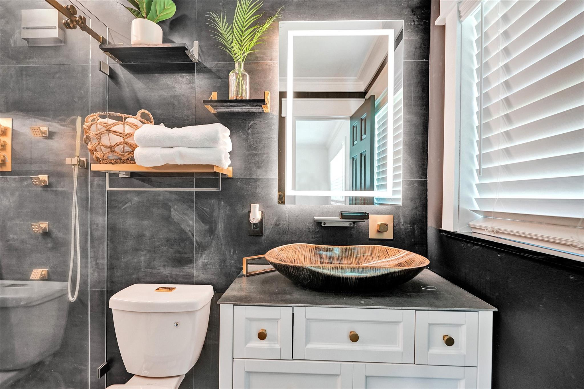 Modern bathroom with dark tile, glass shower, illuminated mirror, and a wood-grain vessel sink.