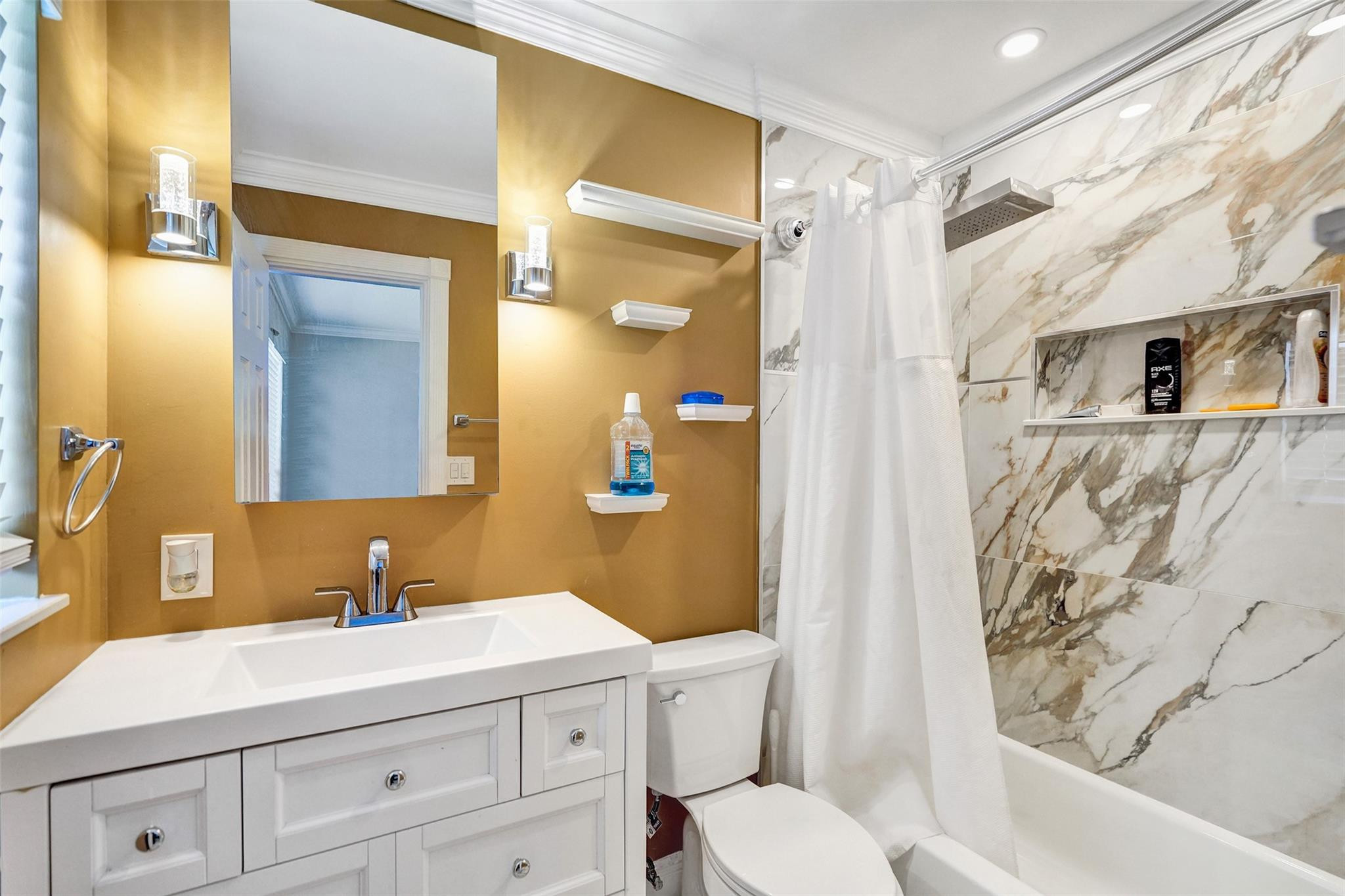 Bathroom with a white vanity, gold walls, and a marble-look tiled tub and shower.