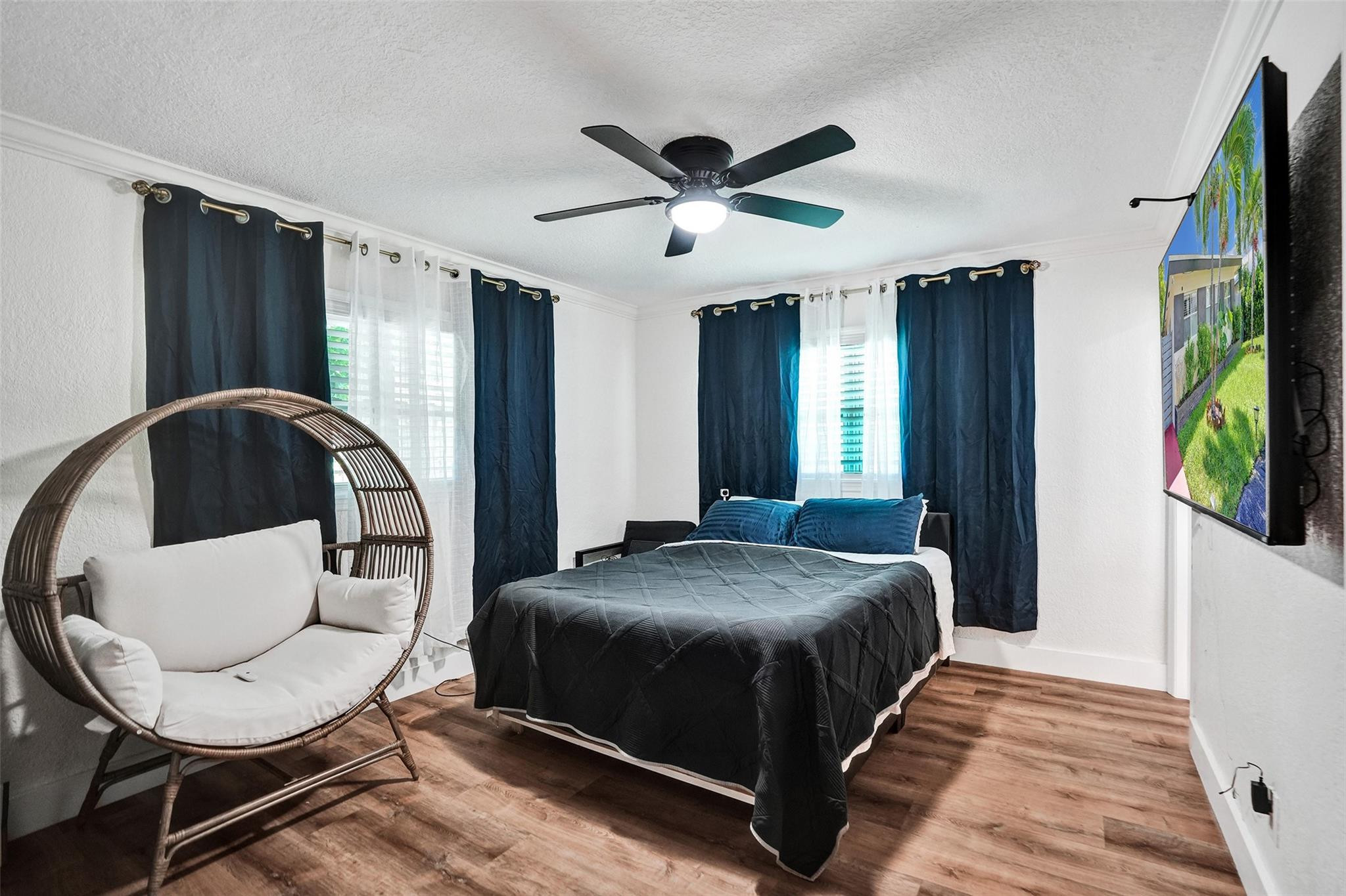 Bedroom with a dark-covered bed, navy curtains, wood-look floors, and a round wicker chair.