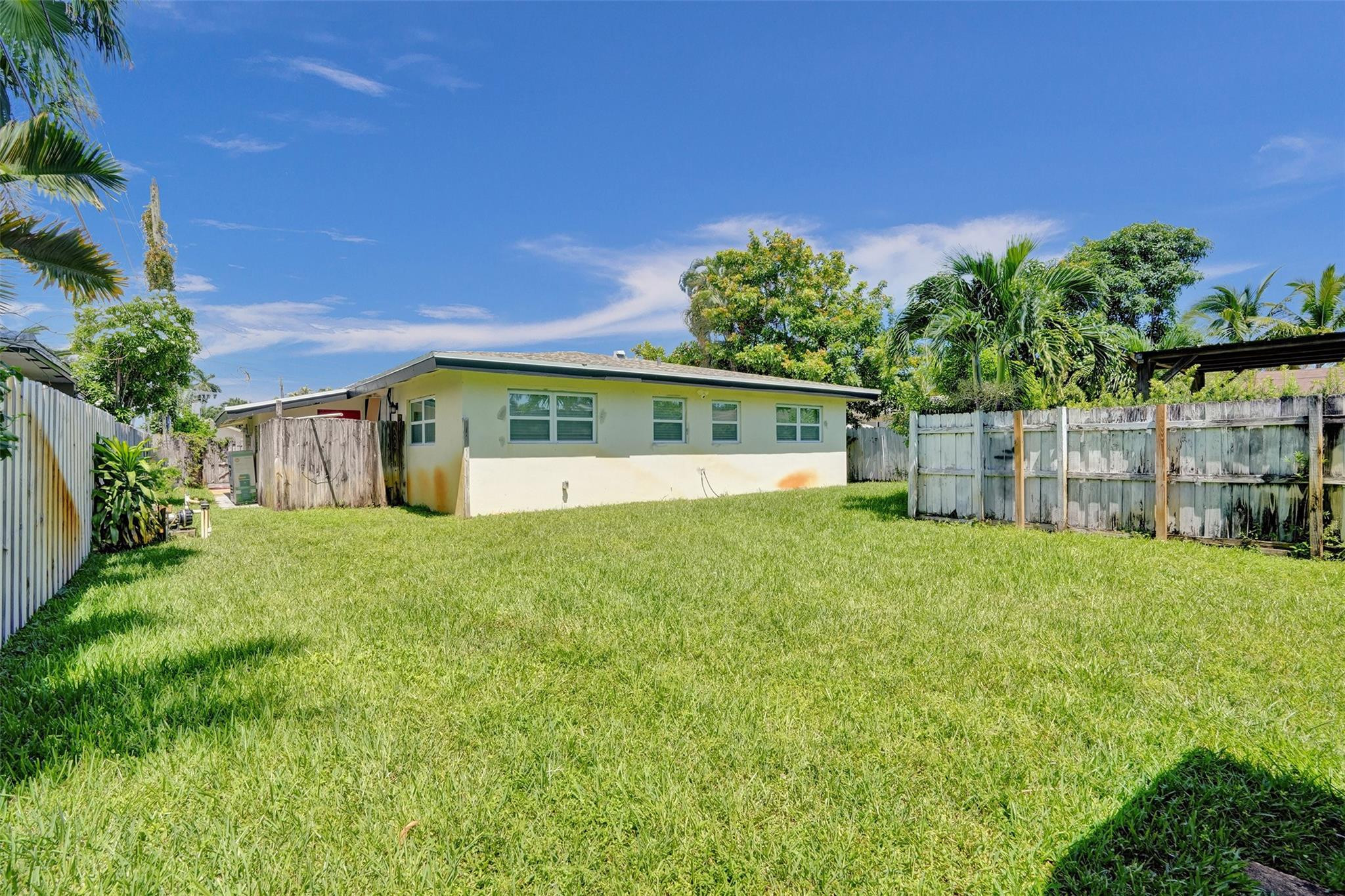 Single-story home behind a broad lawn and weathered privacy fence, with palms and trees around the yard.