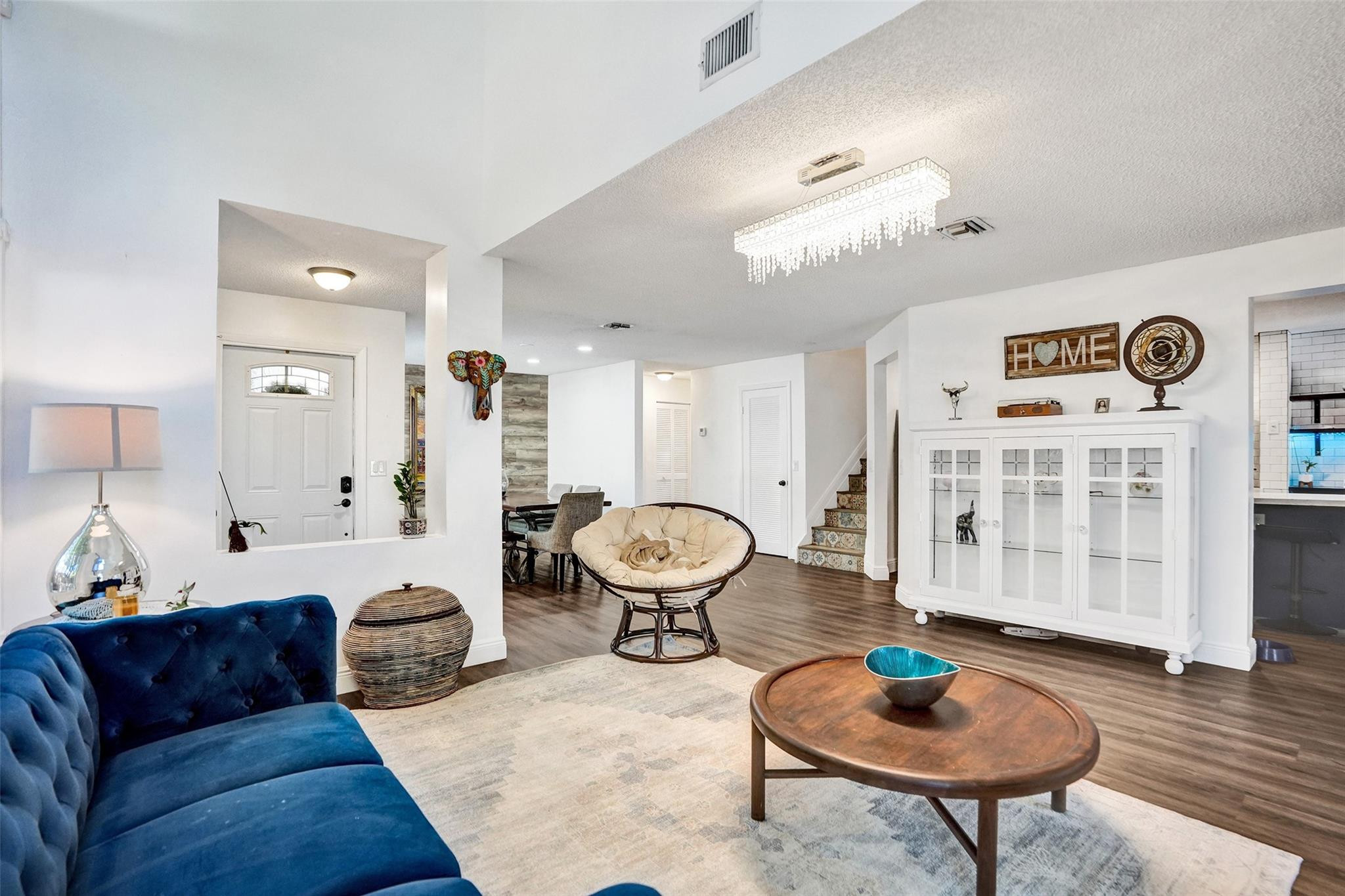 Furnished living room with blue velvet sofa, crystal chandelier, and open dining area.