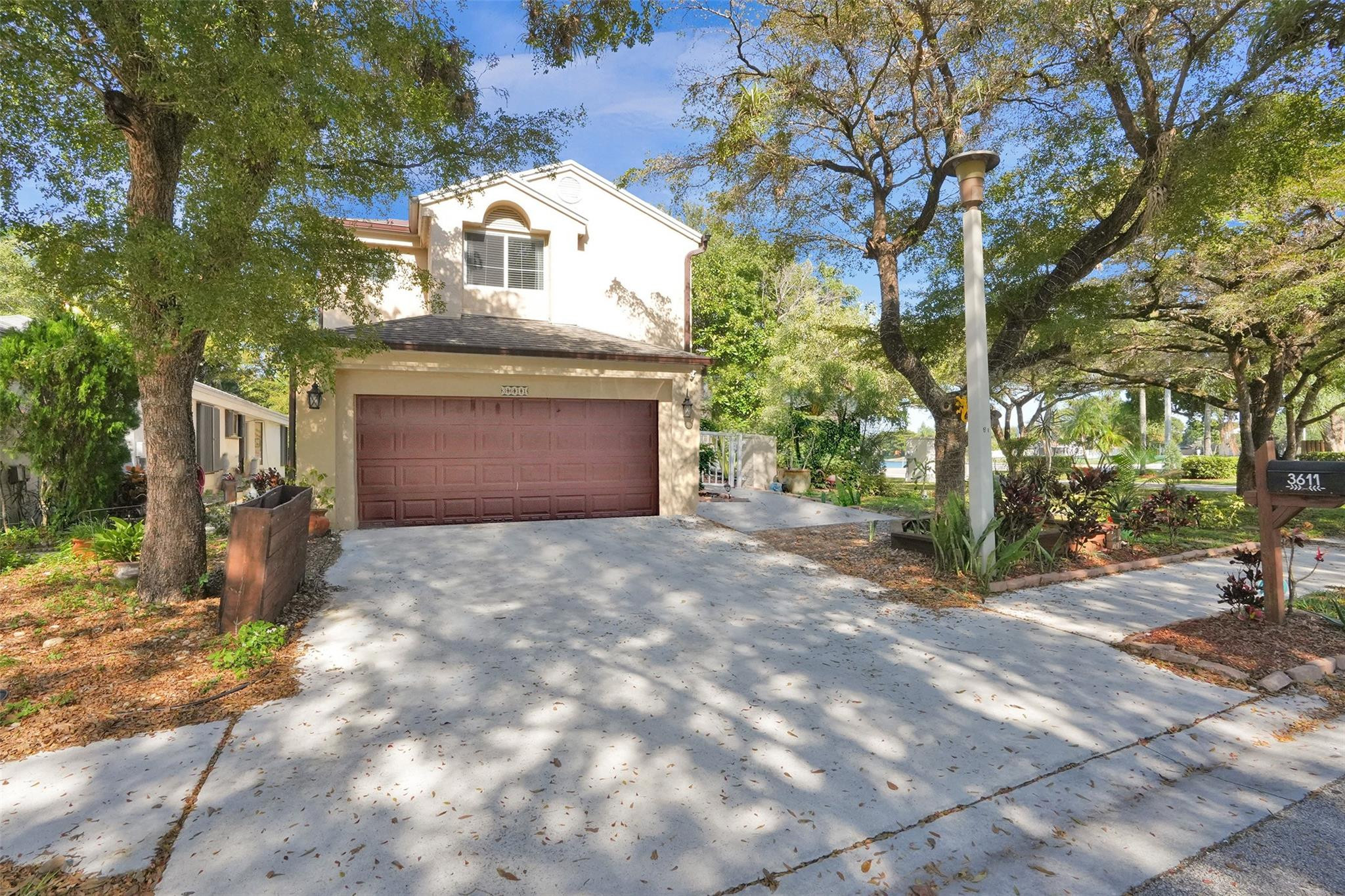 Two-story beige home with a red-brown double garage door, shaded by mature trees.