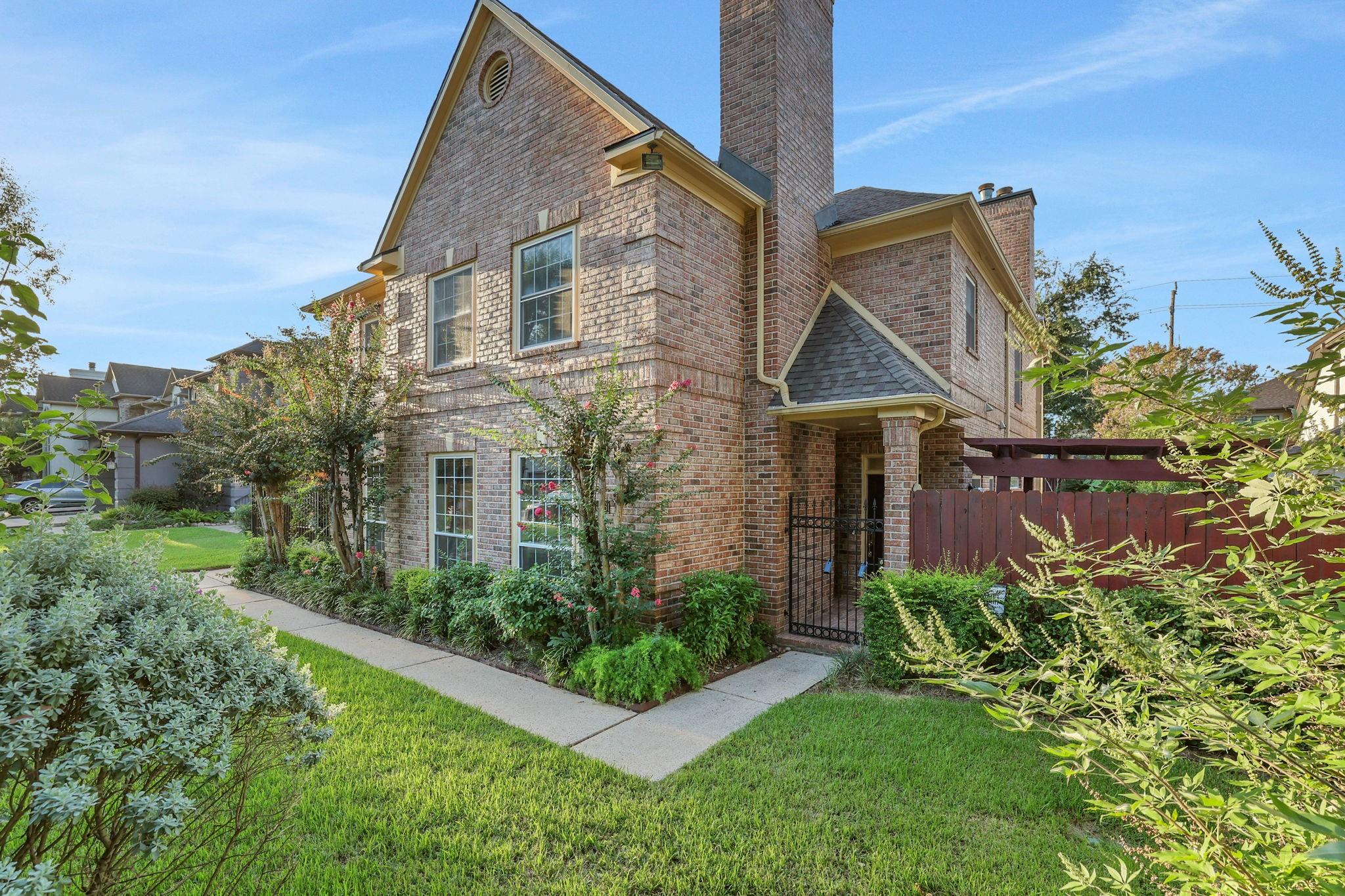 Two-story brick house with a tall chimney, landscaped garden, and gated side entry.