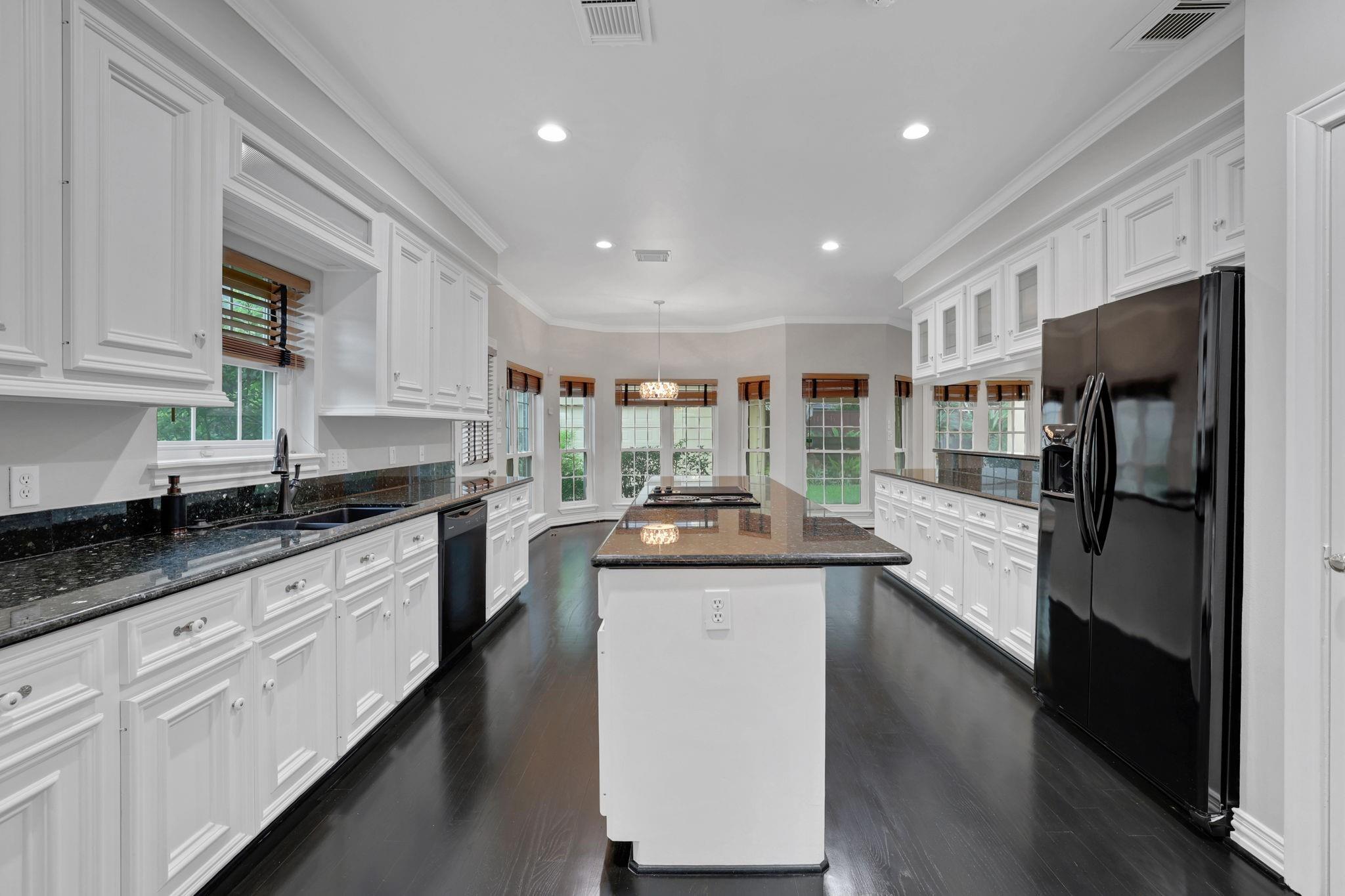 Bright kitchen with white cabinetry, a large granite island, black appliances, and dark hardwood floors.