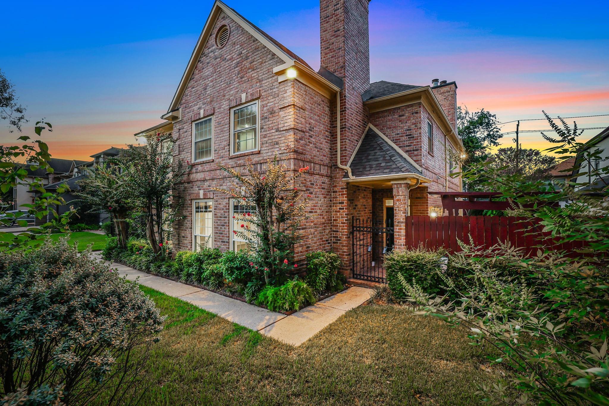 Two-story red brick house with a tall chimney, landscaped garden, and gated entry at dusk.