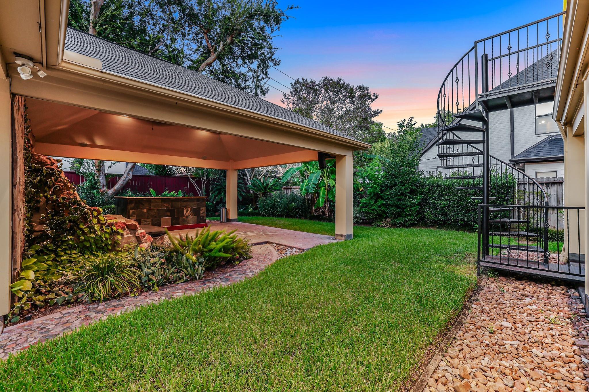Backyard with a covered patio, stone outdoor bar, lawn, and black spiral staircase.
