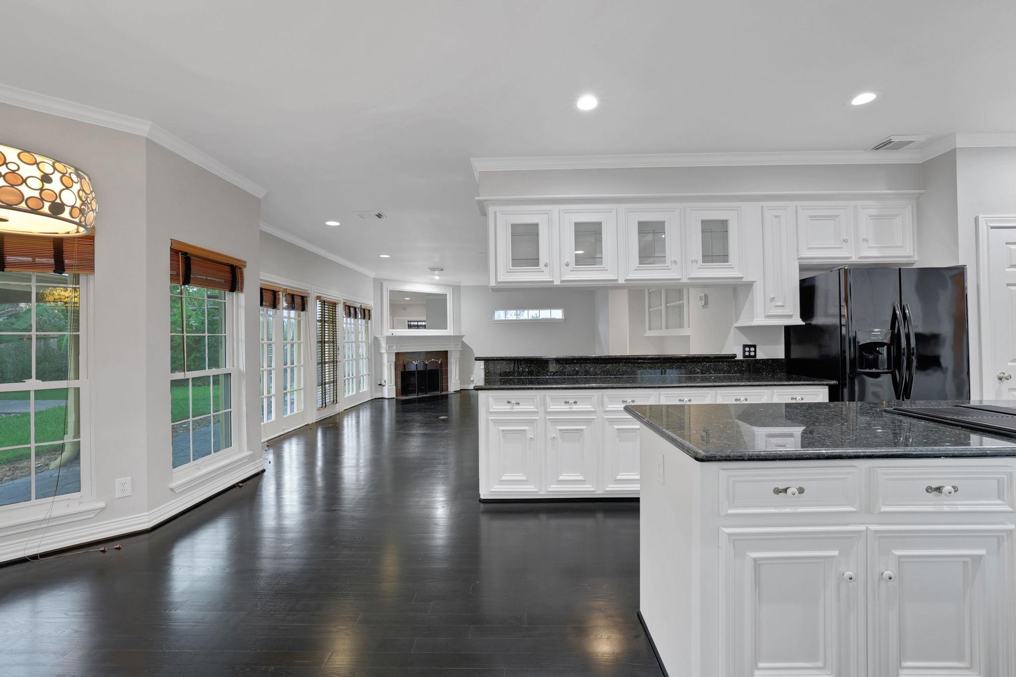 Kitchen with white cabinets, black granite island, dark floors, and tall windows beside a fireplace.