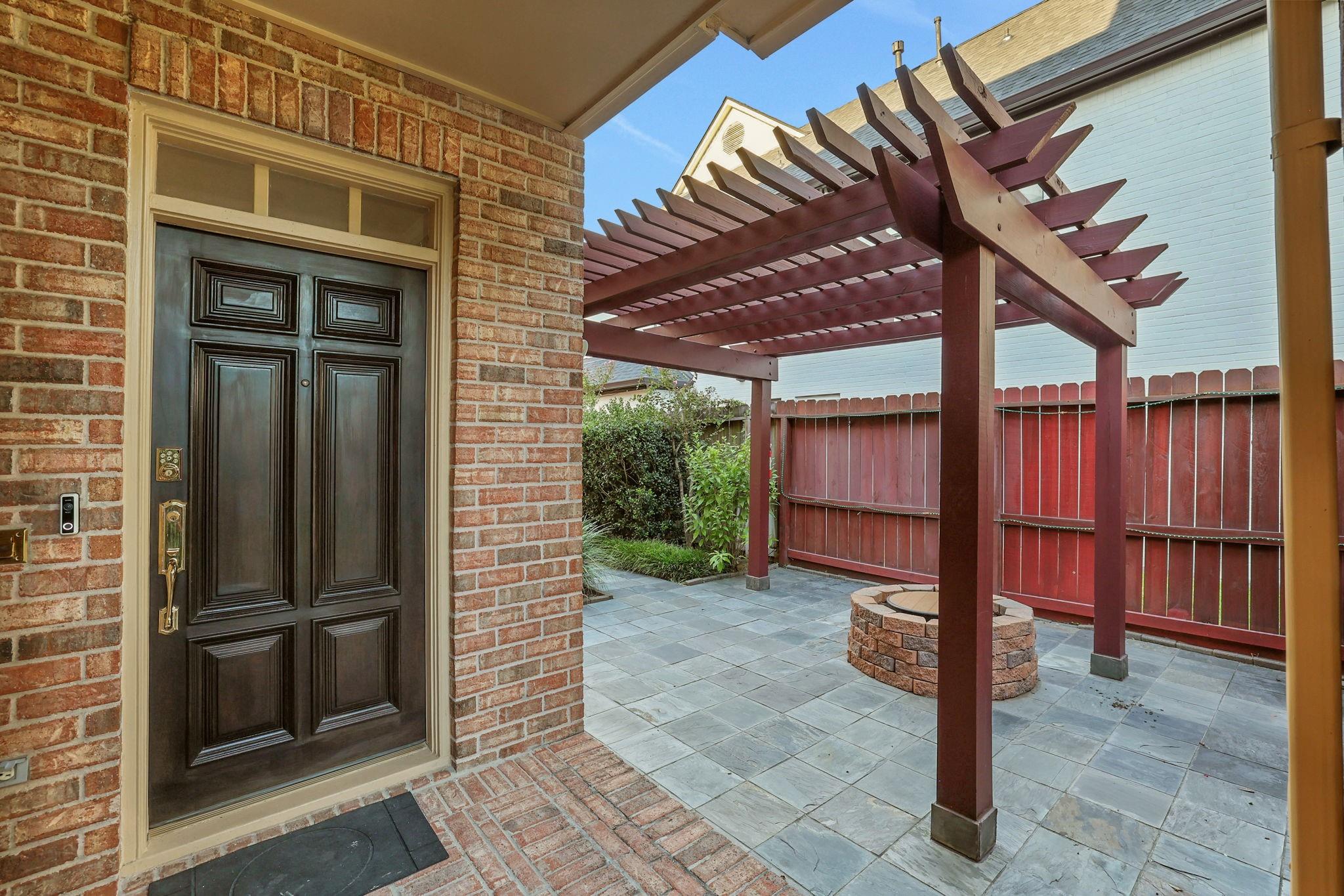 Brick home entrance beside a paved courtyard with a wooden pergola and circular brick fire pit.