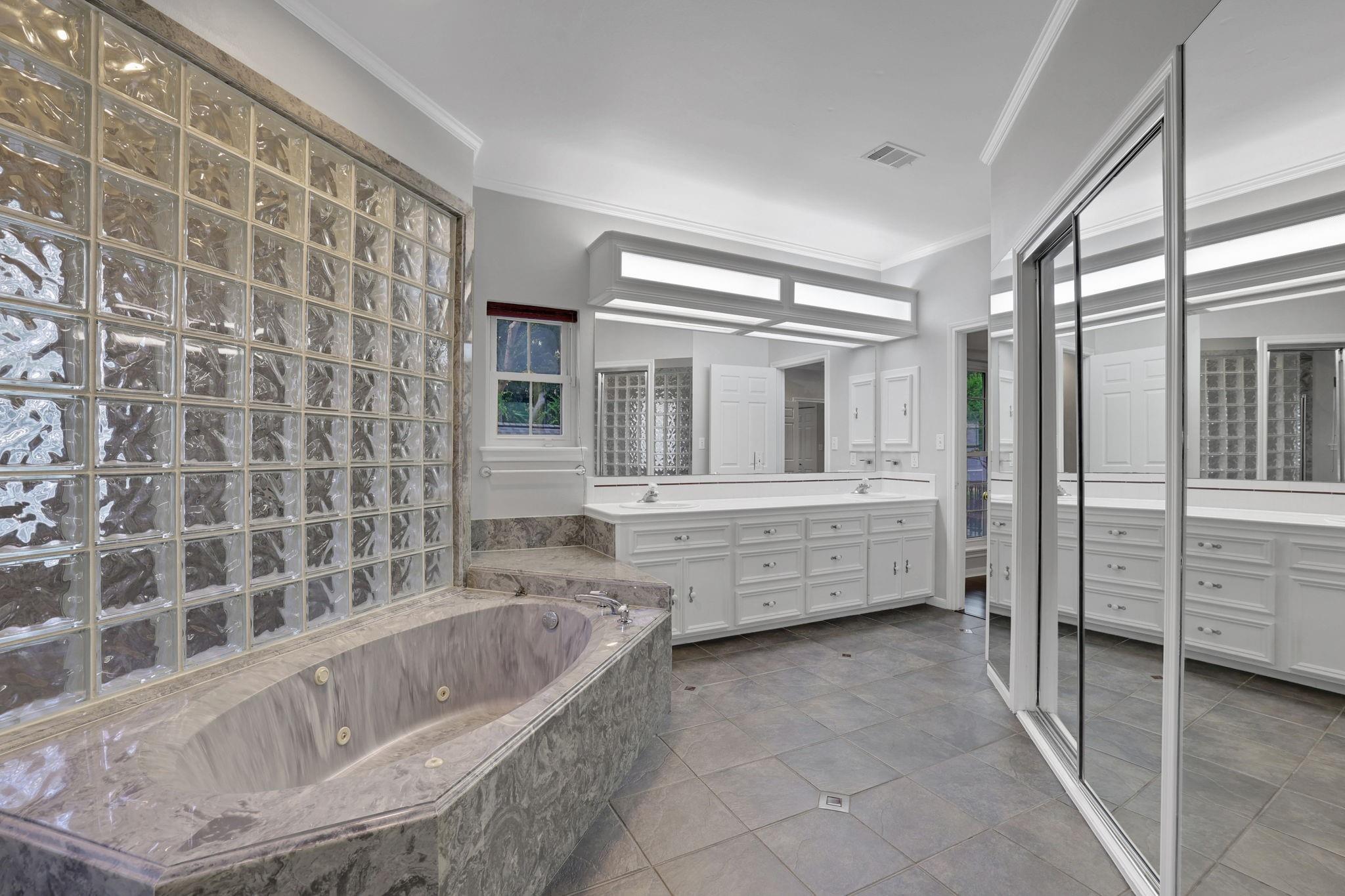Bathroom with a jetted tub, glass-block wall, double vanity, and mirrored closet doors.